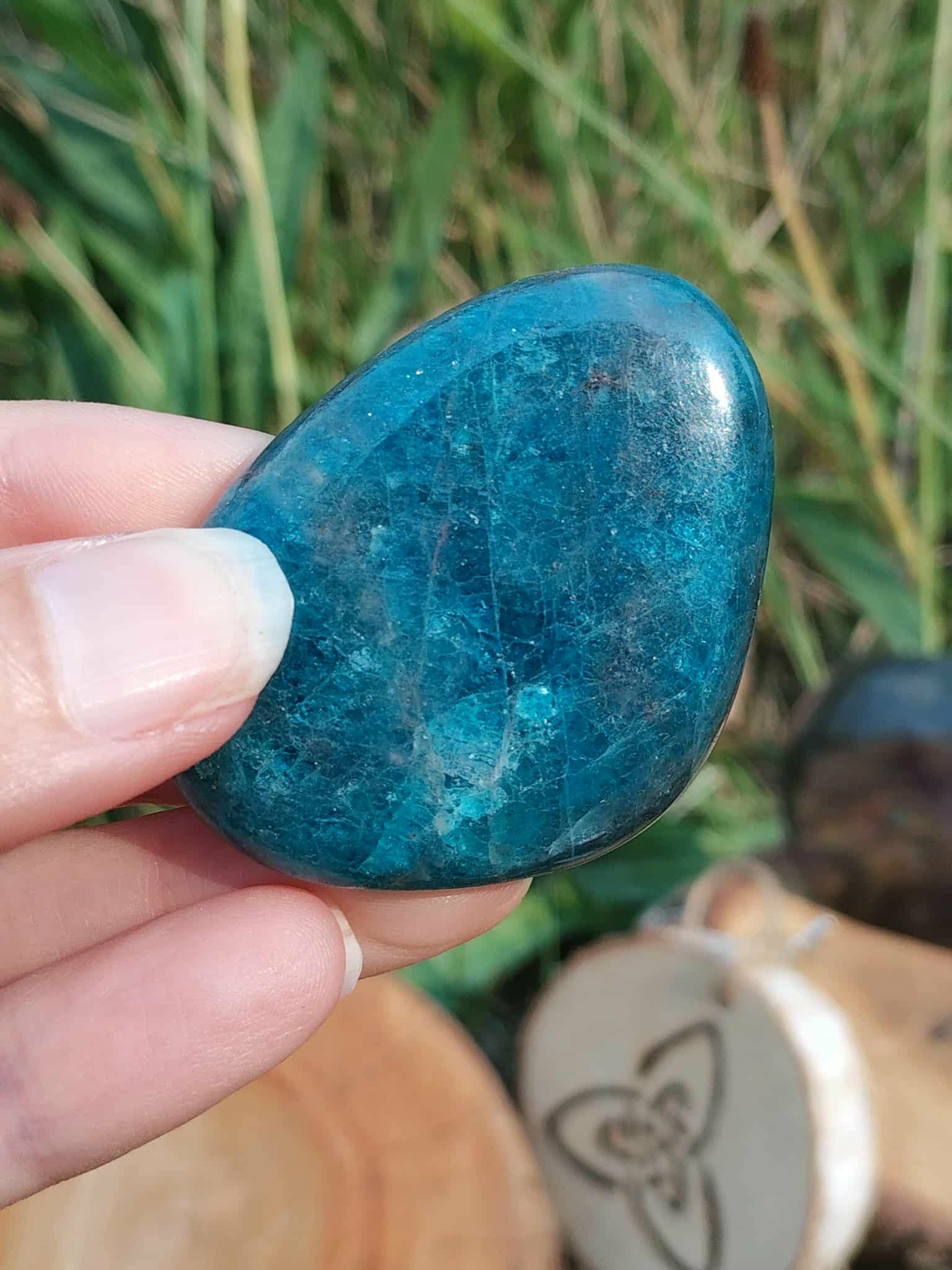 Galet d’apatite bleue poli tenu entre les doigts, avec herbes vertes et décor ésotérique en arrière-plan, pierre naturelle liée au recentrage, à l’authenticité et à la sensibilité apaisée.