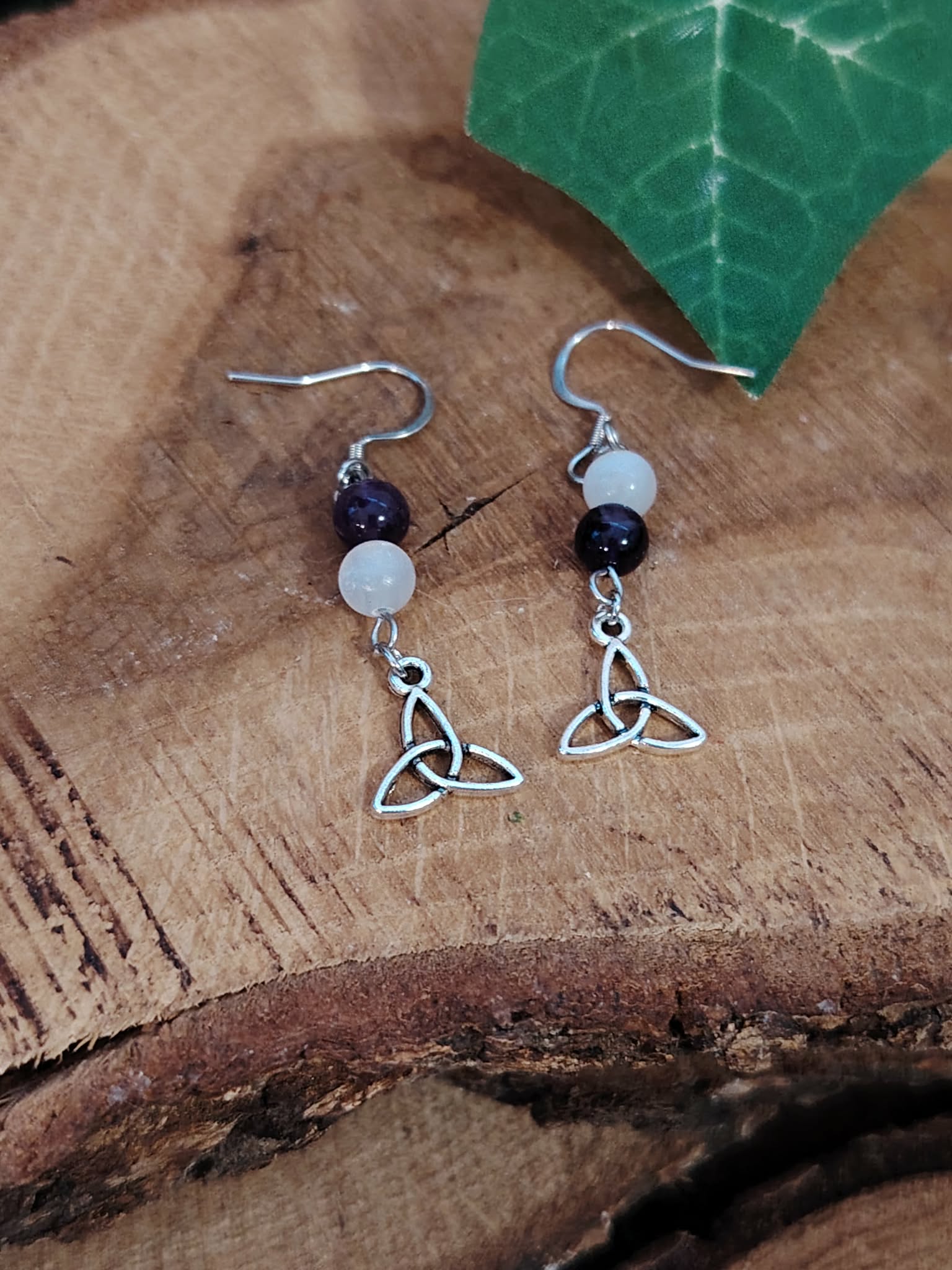 Boucles d’oreilles pendantes triquetra argenté, perles sélénite améthyste posées sur bois avec feuille verte en décor – apporte sérénité, recentrage et intuition – Création Magic Gaïa.