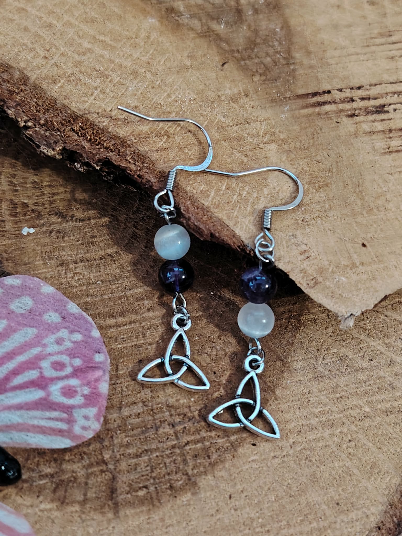 Boucles d’oreilles pendantes triquetra argenté, perles améthyste et sélénite mise en scène sur support bois/minéral – Création Magic Gaïa – invite au calme, à la clarté et à l’intuition.