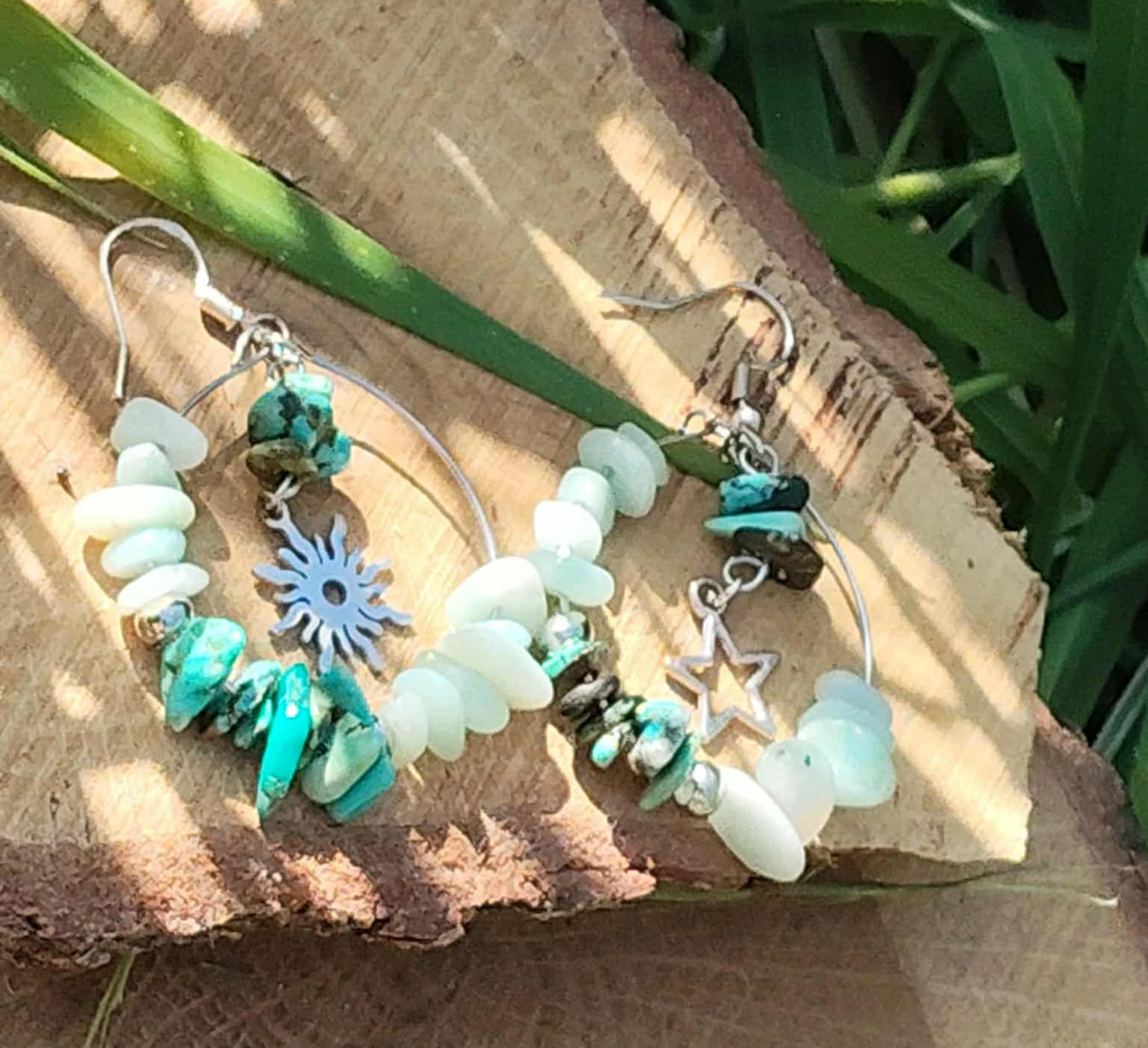 Boucles d’oreilles Ombre &amp; Lumière création Magic Gaïa, anneaux argentés avec amazonite et éclats de turquoise, charms étoile et soleil, photo sur rondin de bois avec feuilles vertes.