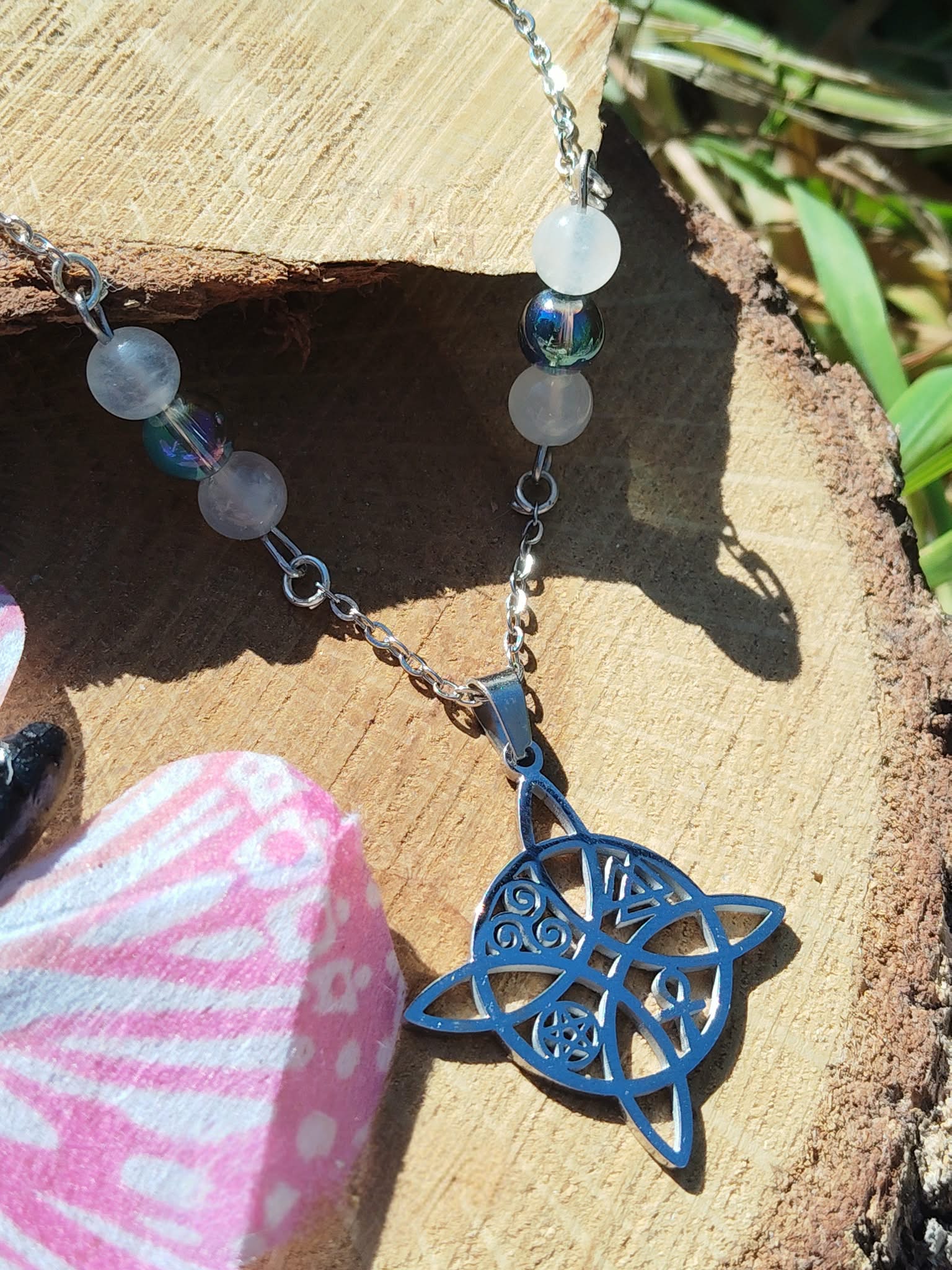 Collier nœud de sorcière bleu posé sur tranche de bois avec tissu rose et feuillage, péristérite et Aqua Aura pour apaisement et énergie protectrice