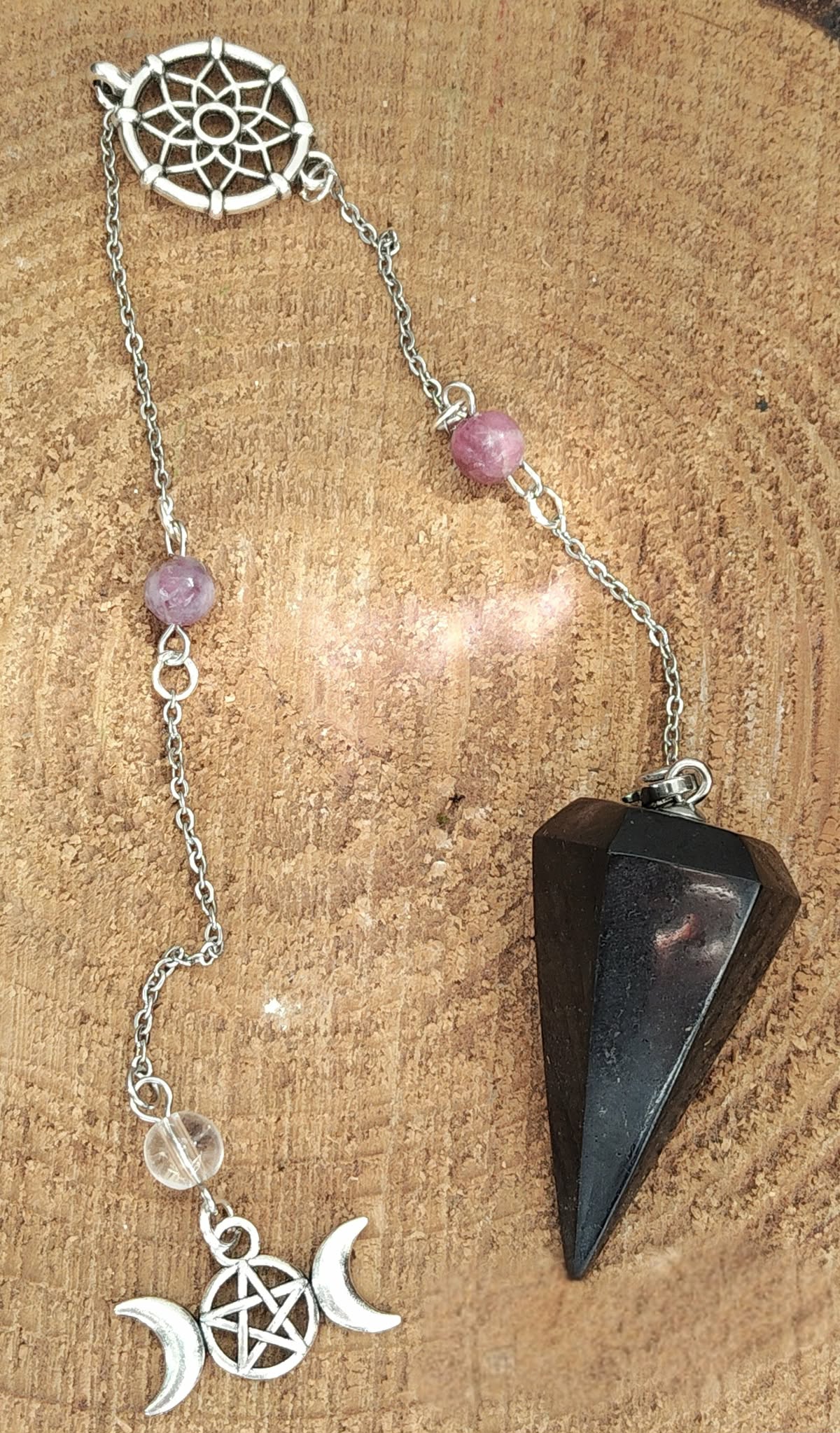 Pendule noir facetté avec chaîne argentée, perles roses et breloques triple lune et attrape-rêves, posé sur bois brut, aide à la protection, à l’intuition et au recentrage.