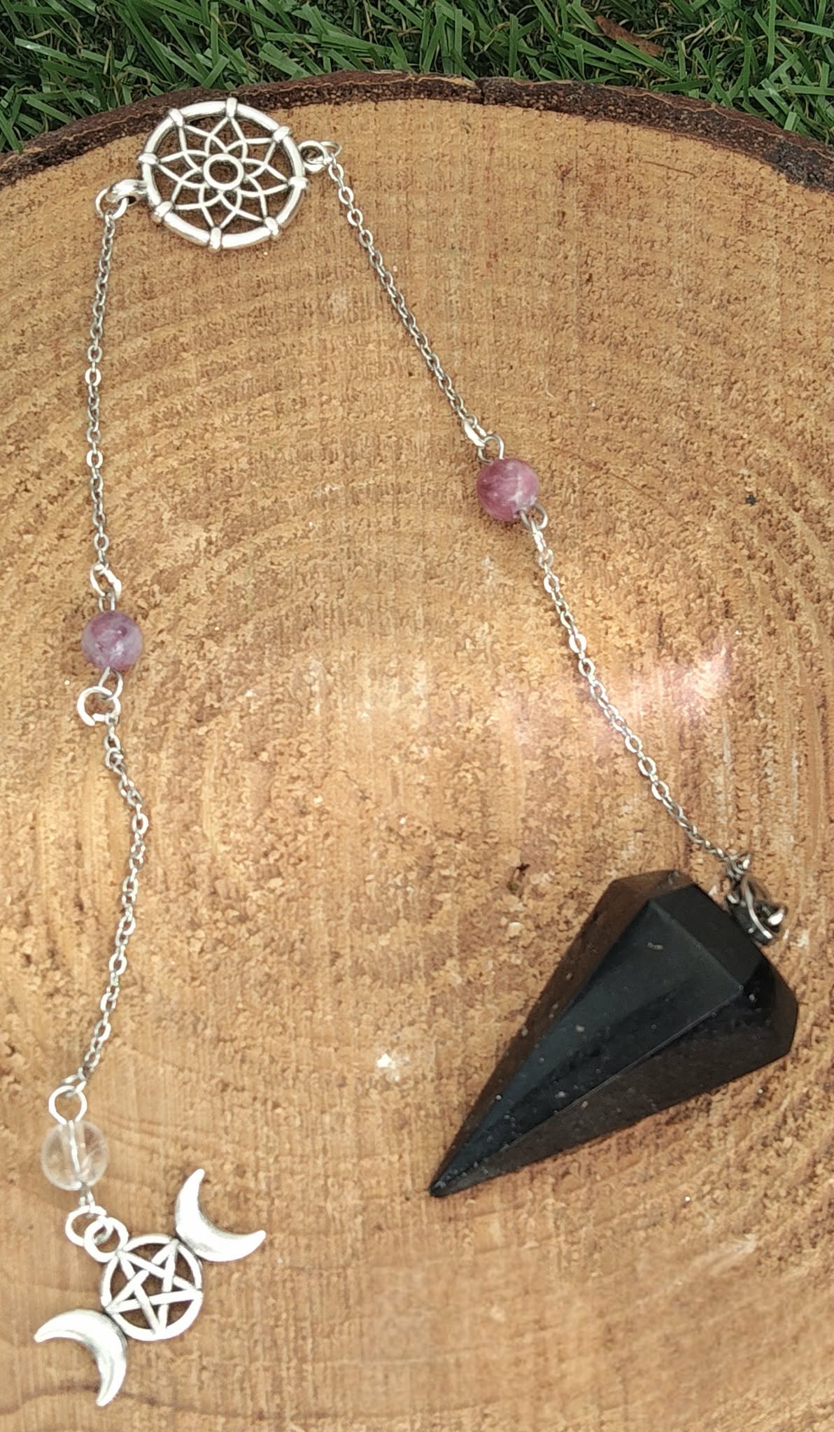 Pendule noir facetté avec chaîne argentée, perles roses et breloques triple lune, posé sur tranche de bois avec bord végétal, apporte protection, ancrage et clarté.