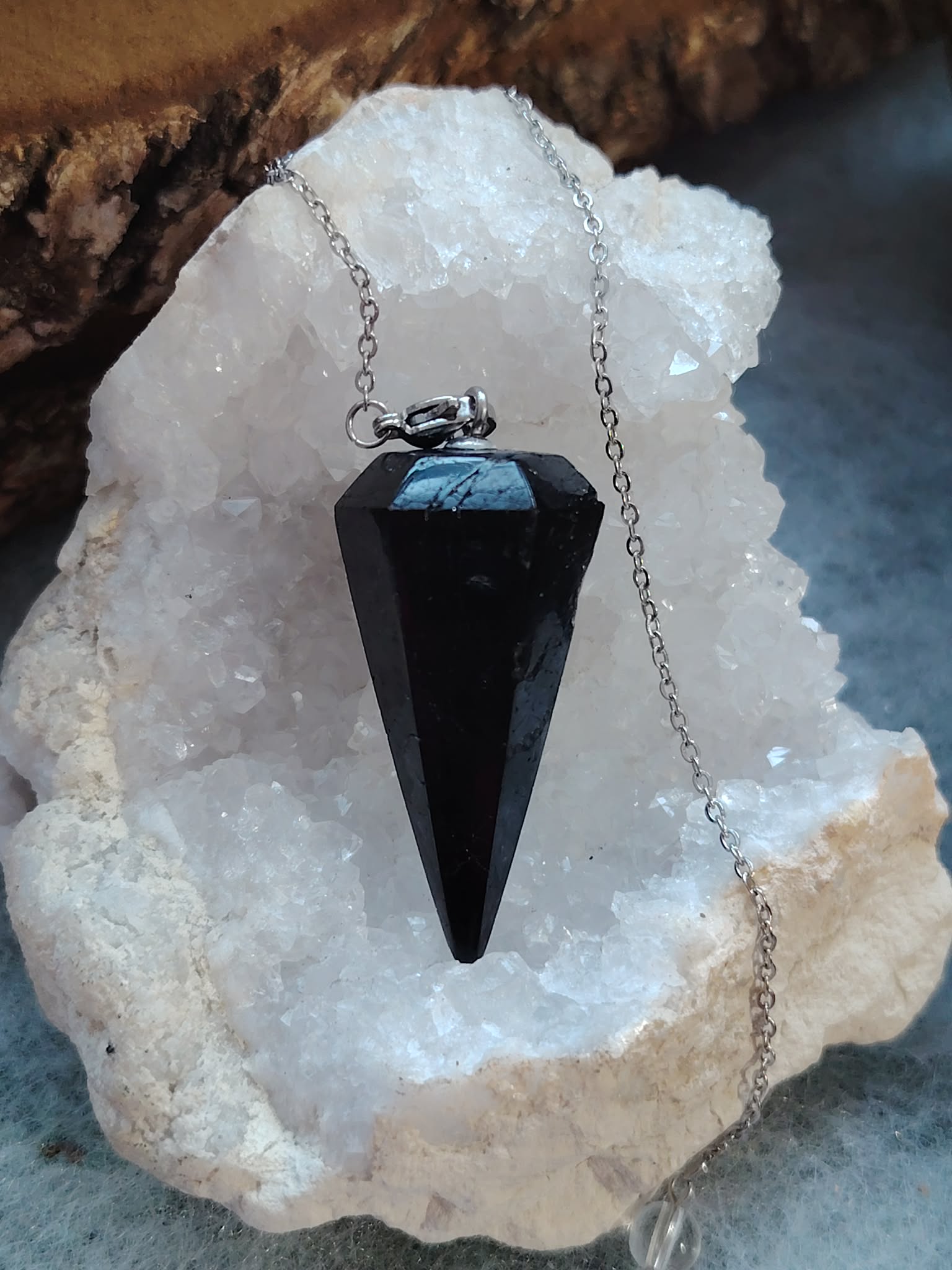 Pendule noir facetté sur chaîne argentée, posé sur un amas de quartz blanc scintillant, apporte protection, ancrage et clarté intérieure.