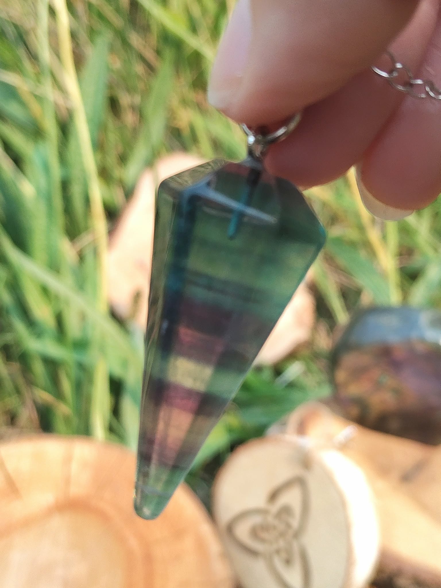 Pendule en fluorite facettée tenu en main, décor herbes vertes et tranche de bois gravée, aide au recentrage, à la clarté et à l’intuition.