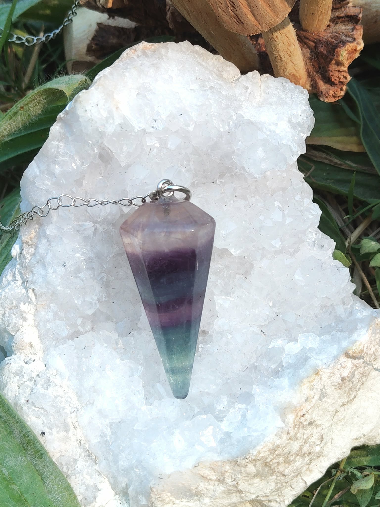 Pendule en fluorite facettée violet/vert sur chaîne argentée, posé sur amas de quartz blanc avec feuillage, aide au recentrage et à la clarté mentale.