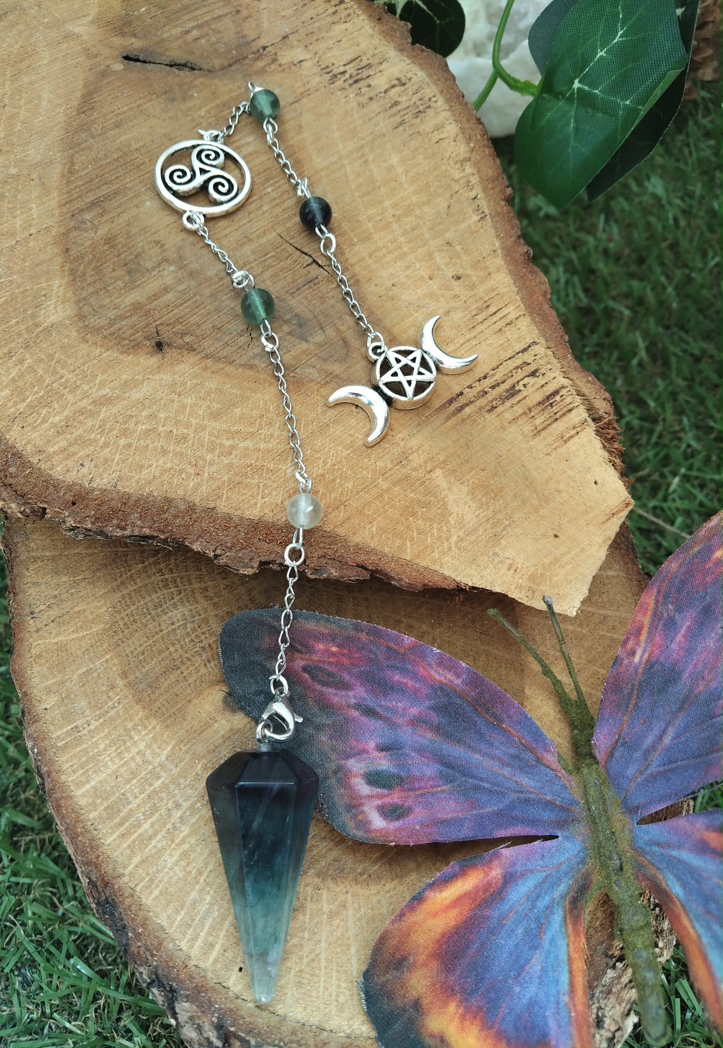 Pendule personnalisé en fluorite multicolore sur chaîne argentée avec breloques ésotériques, posé sur bois naturel avec mousse verte et papillon décoratif, apporte clarté, apaisement et intuition.