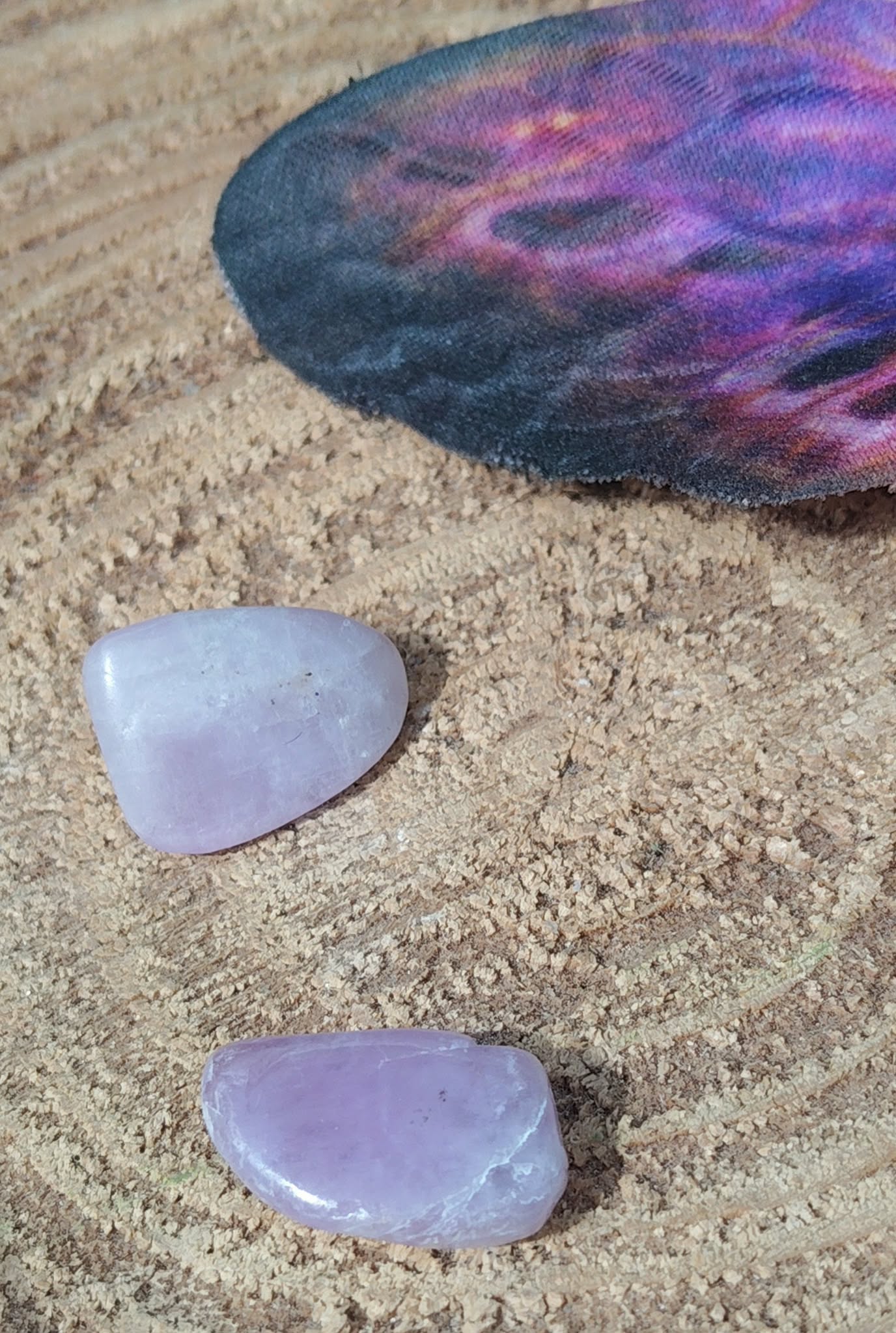 Deux pierres roulées de kunzite rose pâle aux reflets laiteux, posées sur support en bois avec une pierre sombre irisée en arrière-plan, pierres naturelles associées au réconfort, à la paix intérieure et à la sensibilité apaisée.