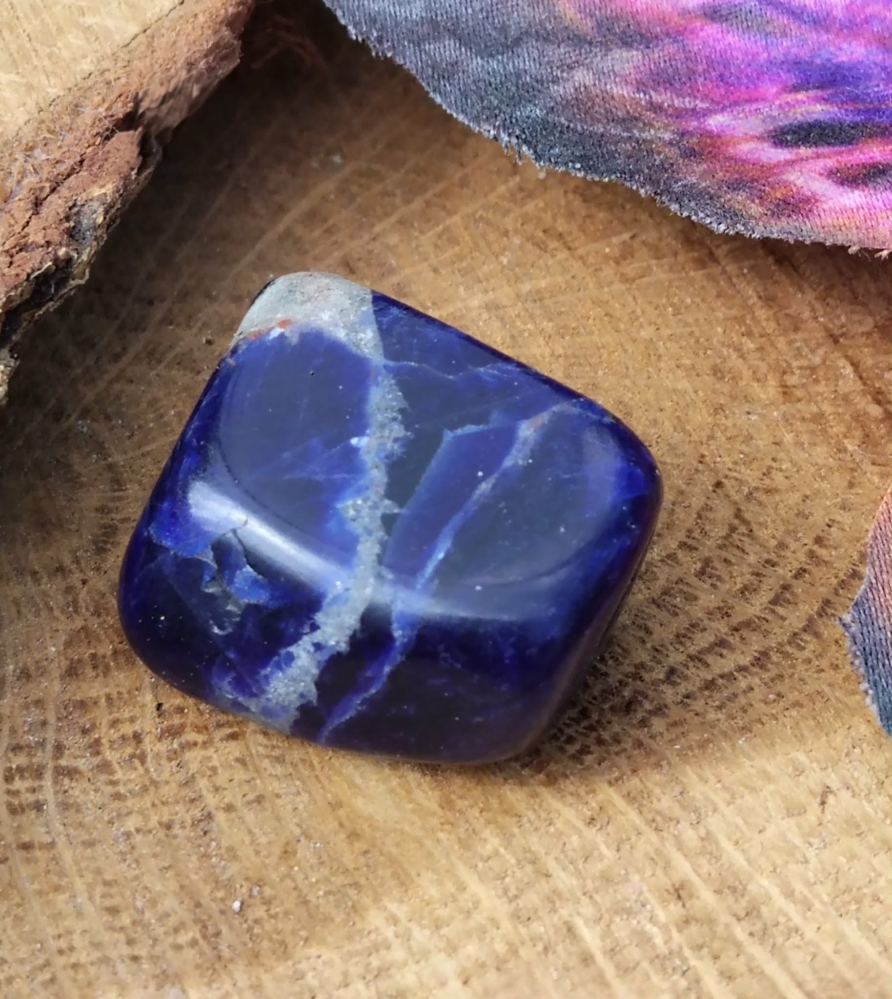 Pierre roulée de sodalite bleu nuit aux veines blanches, galet poli carré posé sur tranche de bois en décor naturel, pierre associée à la concentration paisible, à l’assurance et à la parole juste.