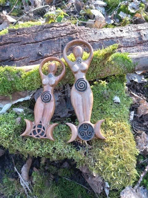 Duo de statuettes Gaïa en bois style wicca, spirale gravée et triquetra sur base triple lune, posées sur mousse et écorce en décor forêt; évoque ancrage, harmonie, protection douce, énergie féminine et connexion à la nature.