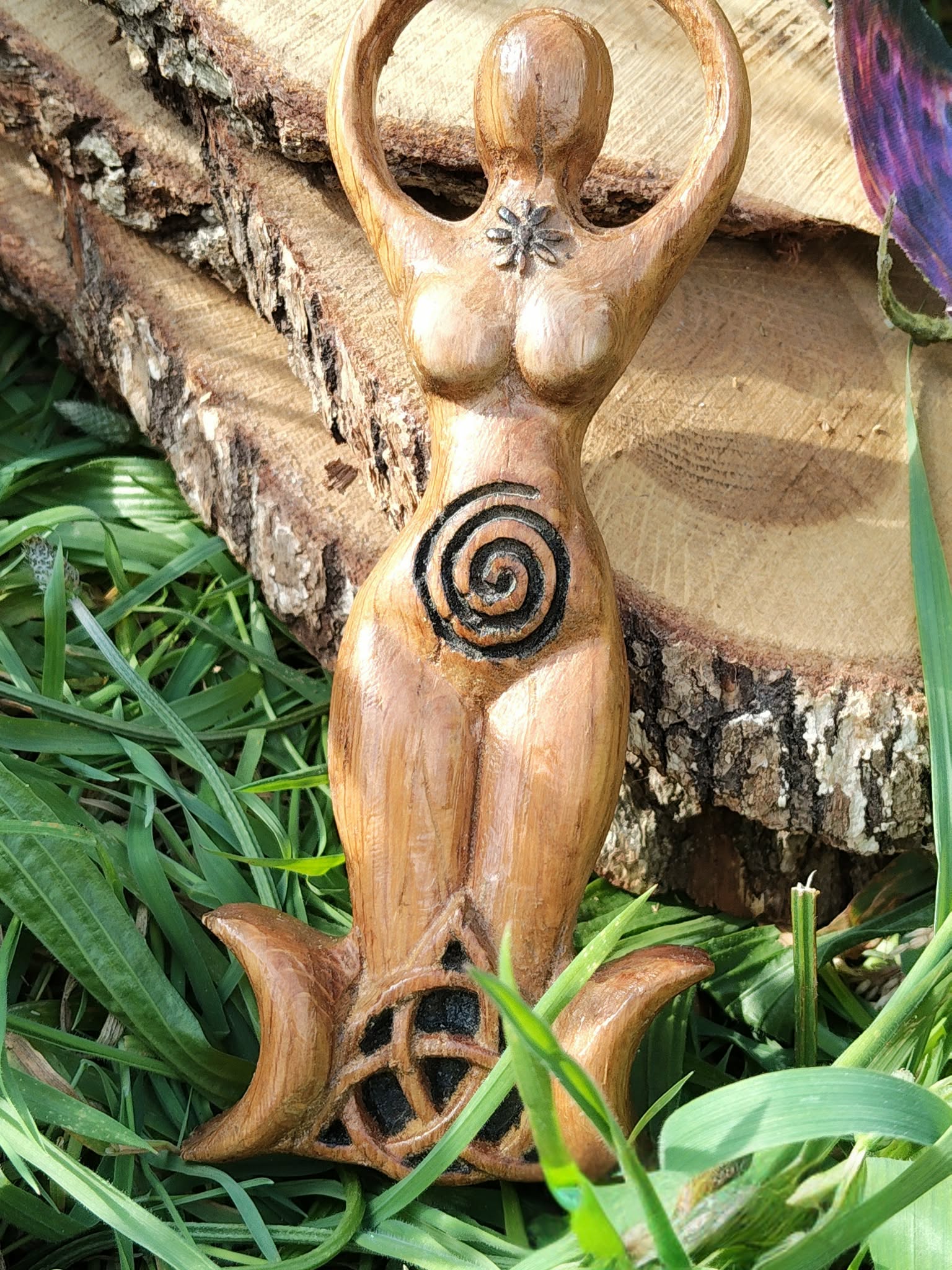 Statuette Gaïa déesse en bois avec spirale, posée dans un décor végétal (mousse/plantes) contre une écorce, ambiance nature; inspire ancrage, apaisement, protection douce, intuition et harmonie.