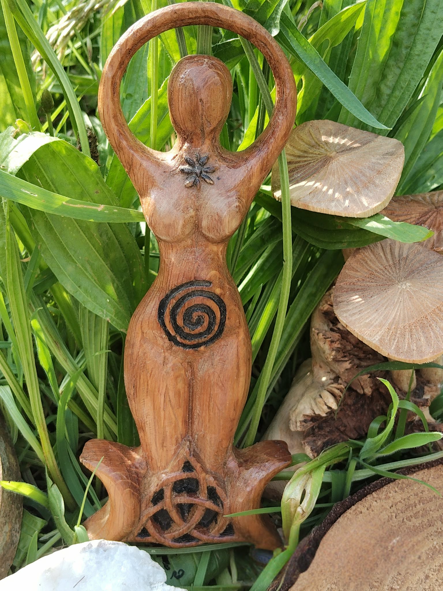 Statuette Gaïa en bois style wicca avec spirale et triquetra, mise en scène dans un décor feuillage avec galets et rondins; suggère ancrage, sérénité, harmonie, protection douce et connexion à la nature.