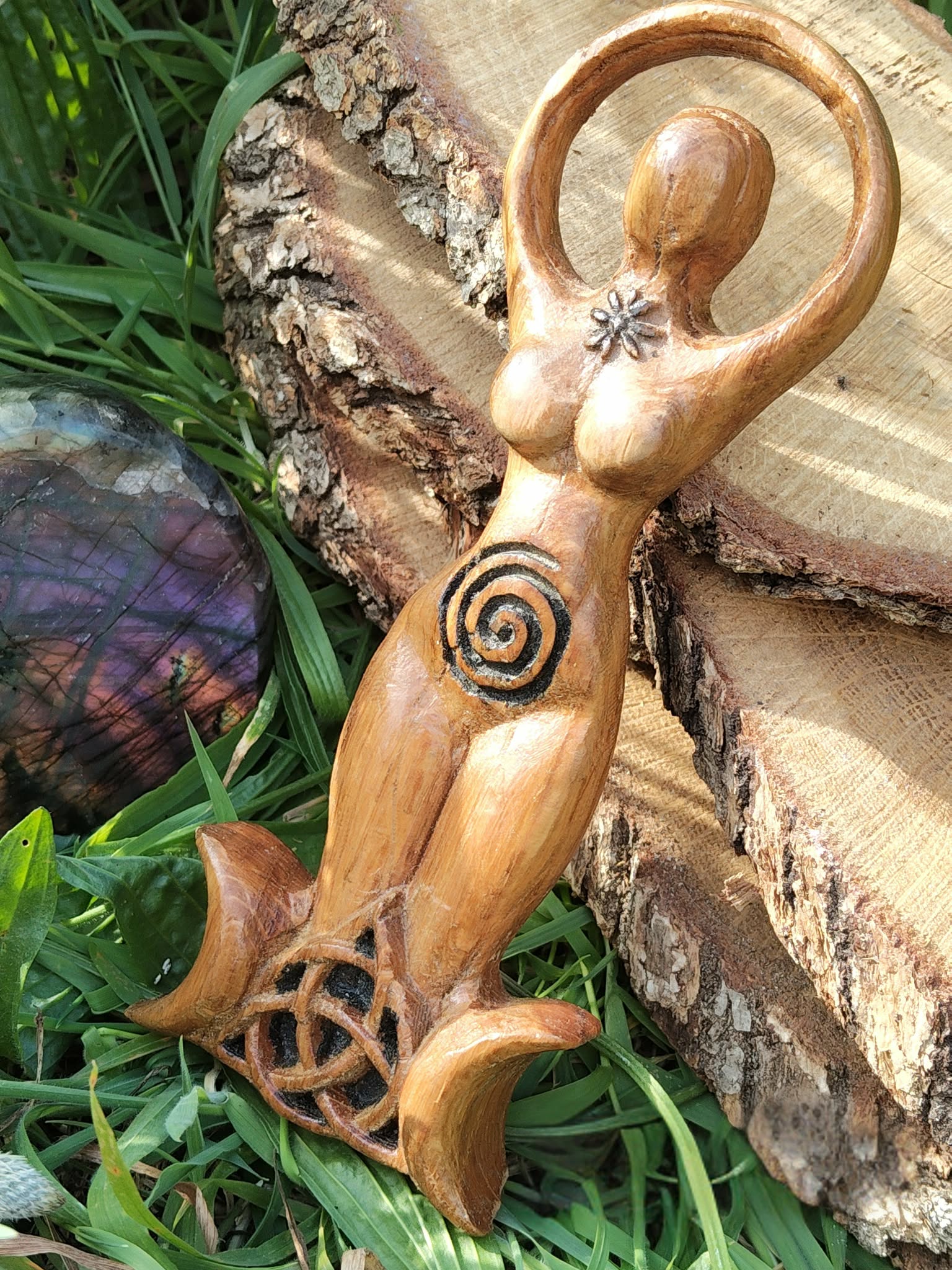 Statuette Gaïa en bois style wicca, spirale gravée et triquetra sur base triple lune, posée contre une écorce avec feuillage et pierre violette; évoque enracinement, protection, équilibre, intuition et lien à la Terre.