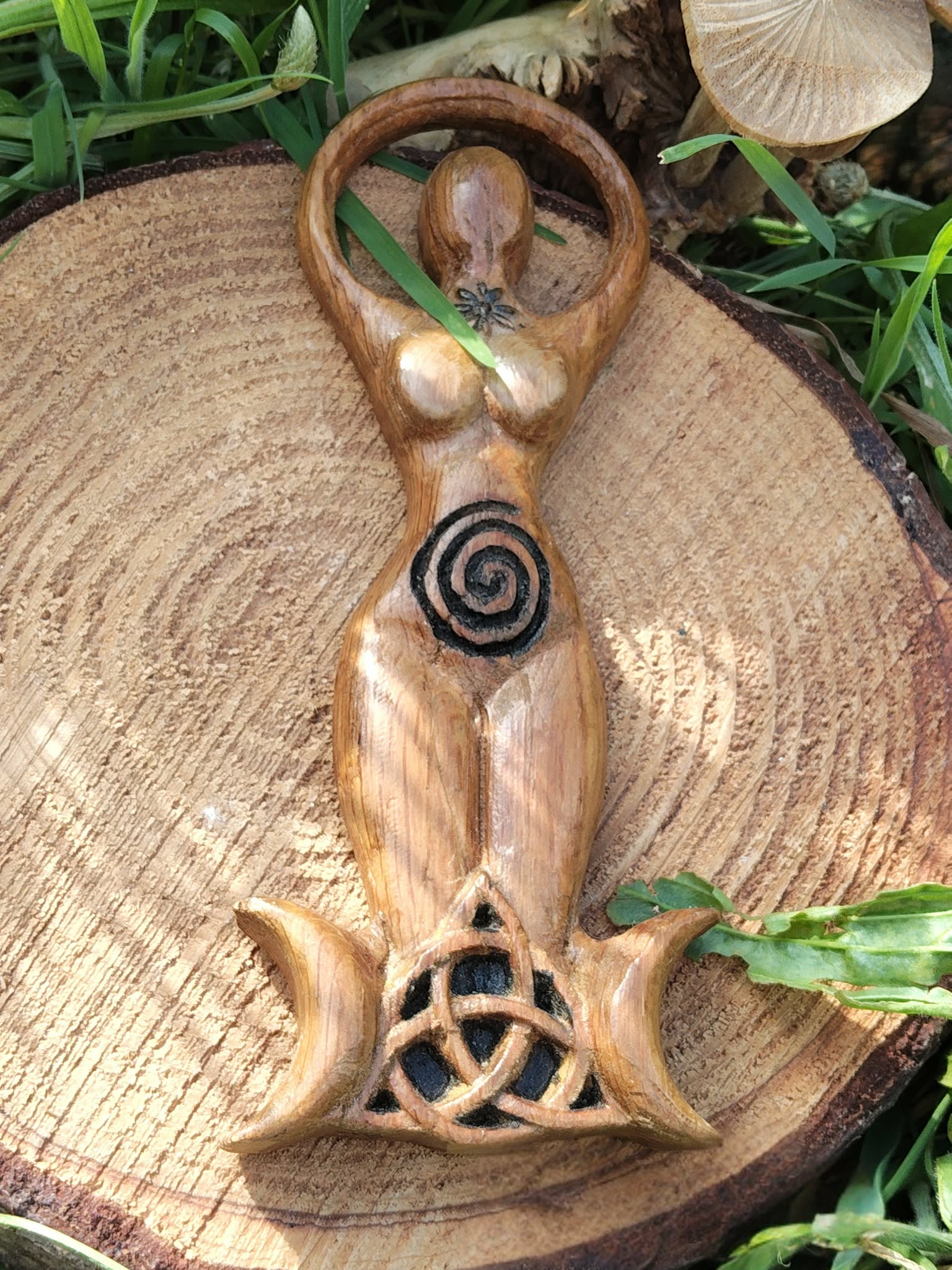 Statuette Gaïa (déesse) en bois style wicca avec spirale et triquetra, posée sur une tranche de bois dans un décor végétal; évoque ancrage, harmonie, protection douce, intuition et énergie féminine, idéale pour autel et déco ésotérique.