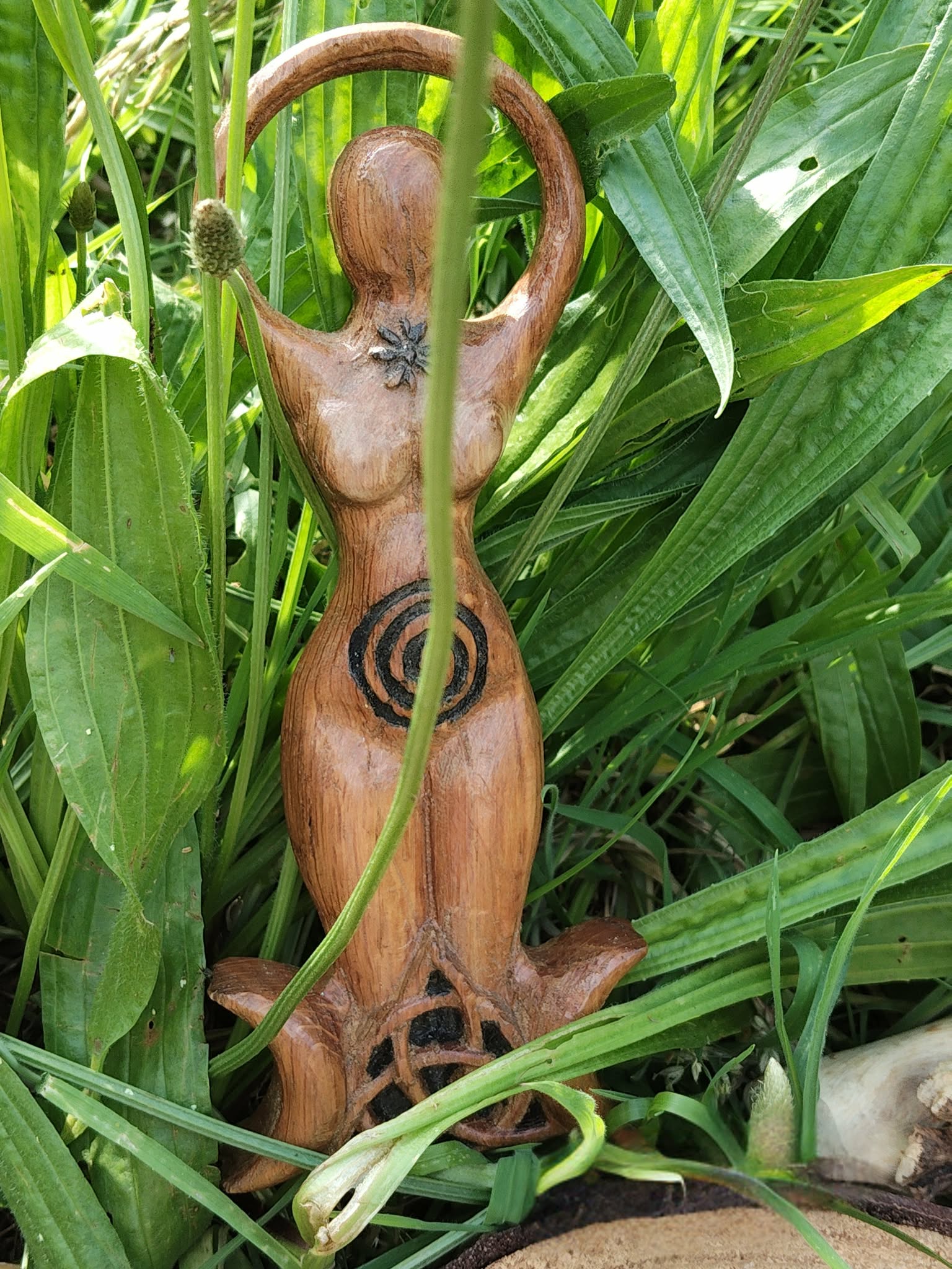 Statuette Gaïa en bois style wicca avec spirale et triquetra, photographiée au cœur des feuilles vertes dans un décor nature; invite à l’ancrage, au calme intérieur, à l’harmonie, à la protection douce et à l’intuition.