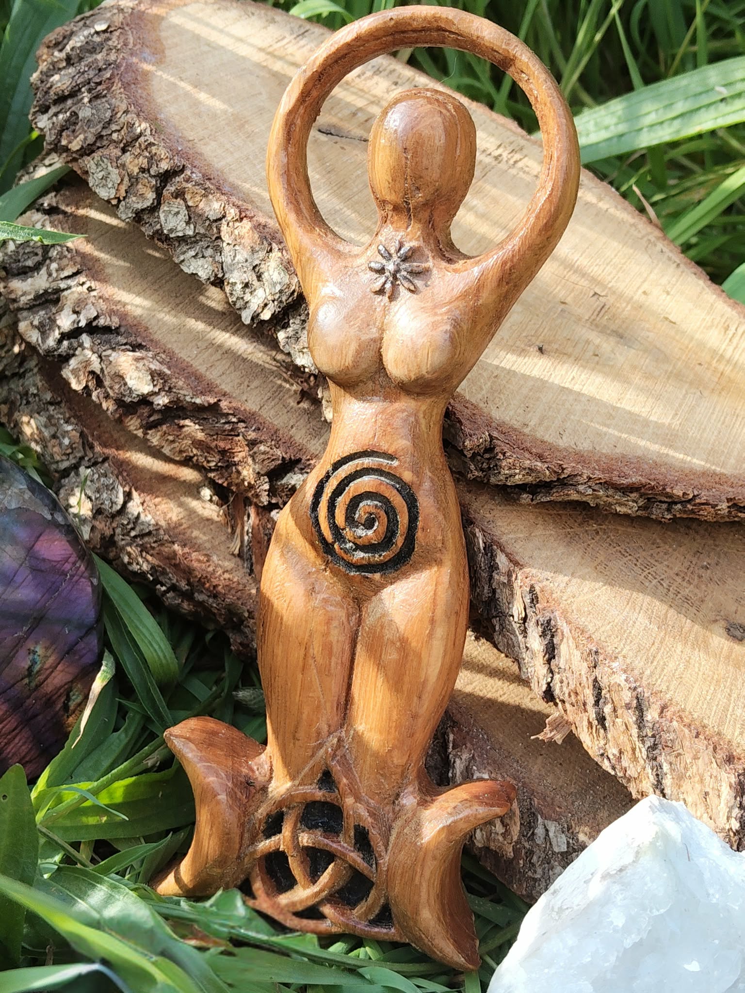 Statuette Gaïa en bois style wicca, spirale au ventre et triquetra sur base triple lune, mise en scène nature avec écorce, pierres et feuillage; inspire ancrage, protection, harmonie, intuition et énergie féminine sacrée.
