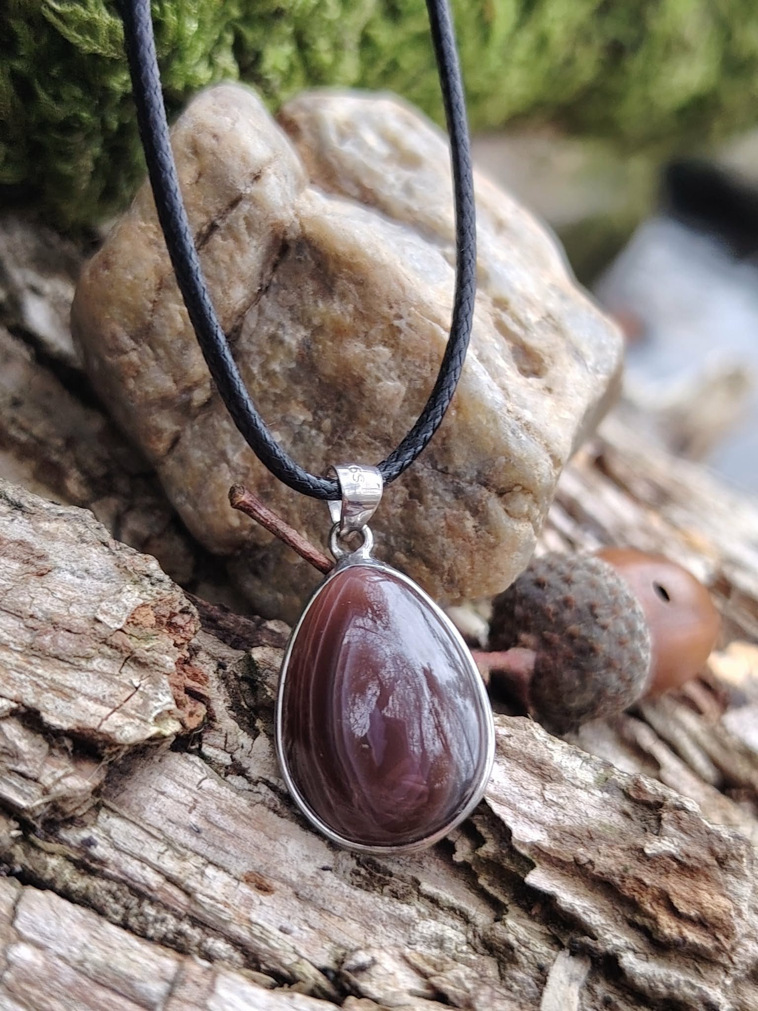 Pendentif agate du Botswana ovale sur cordon noir, bandes brun-gris serties métal argenté, photo en forêt en Bretagne sur bois et pierres naturelles