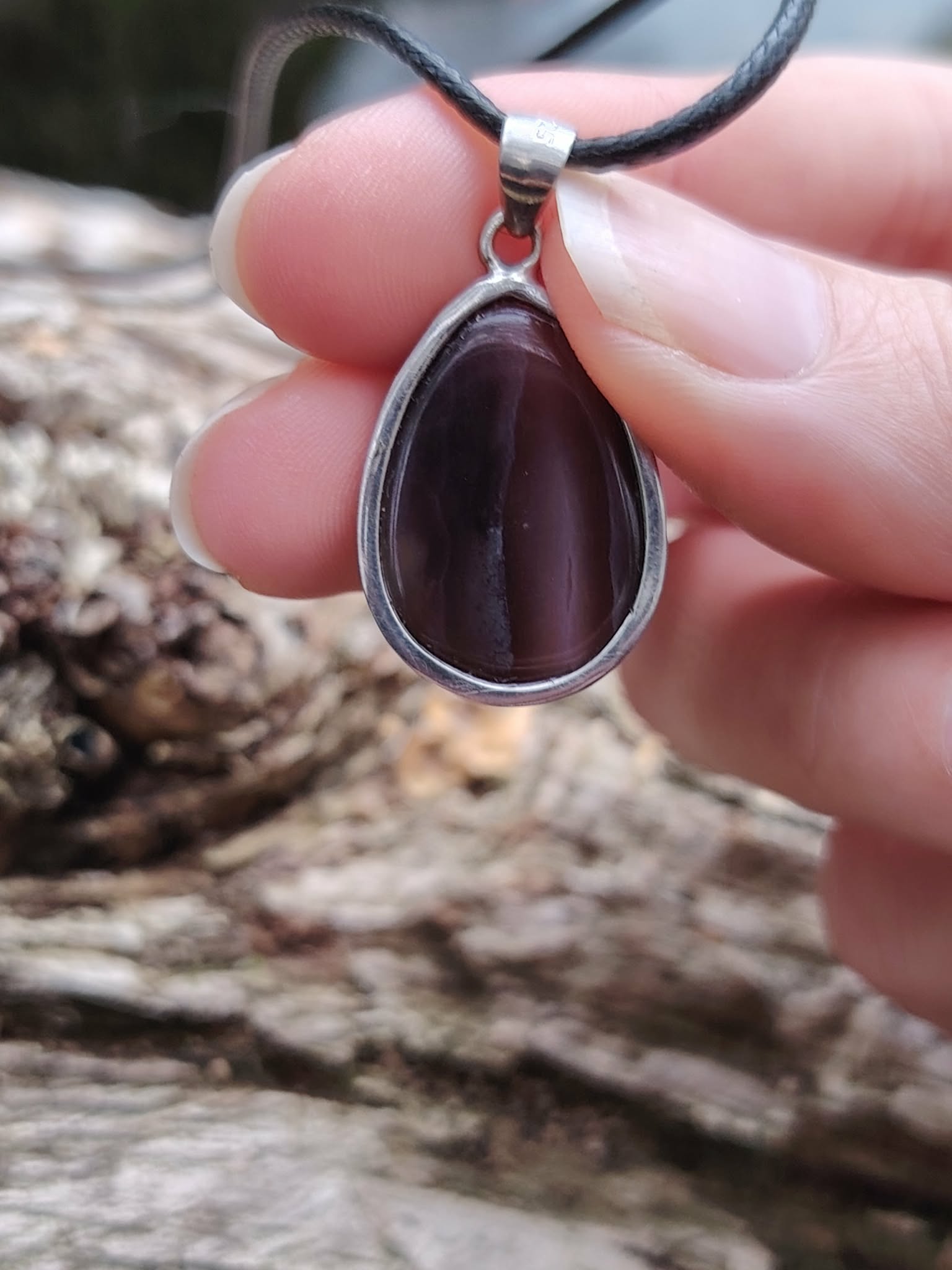 Pendentif agate du Botswana à fines bandes brun-gris sur cordon noir, tenu entre les doigts sur fond de roches, stabilité douce et présence intérieure