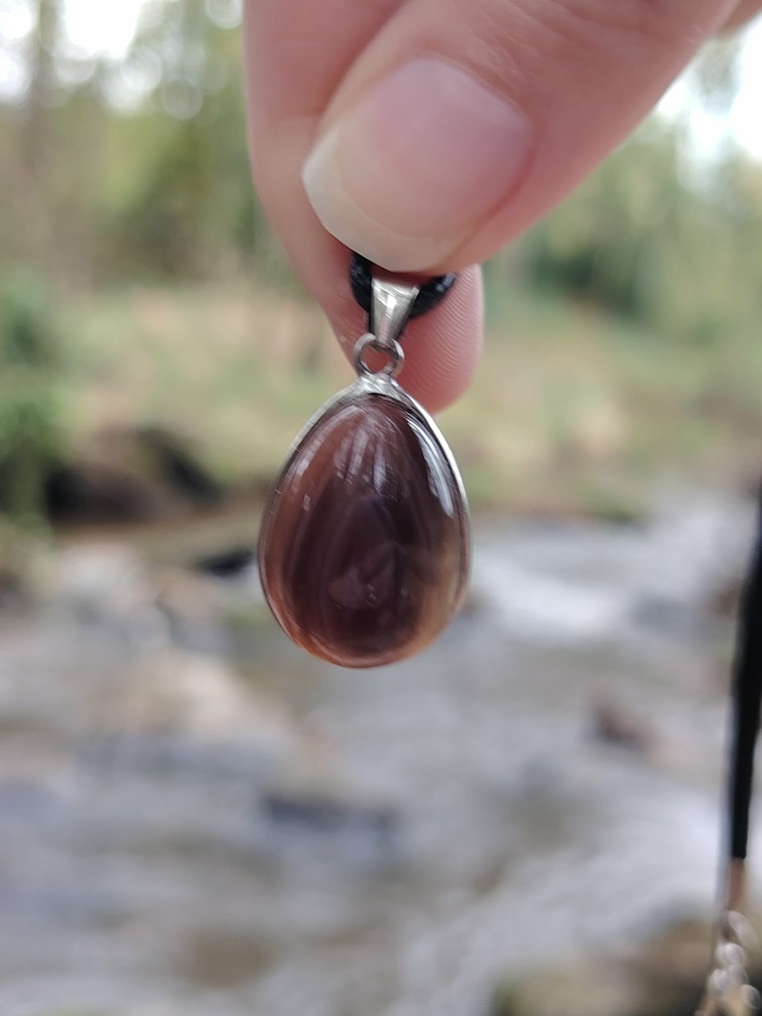 Pendentif agate du Botswana goutte tenu entre les doigts en extérieur sur pierres en Bretagne, bandes brun-gris et énergie de réconfort