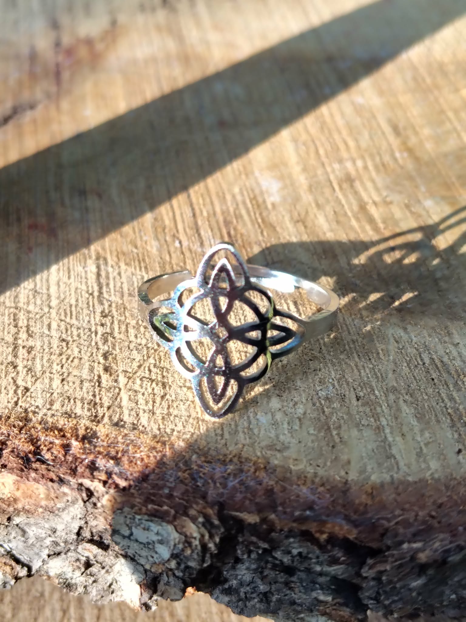 Bague réglable acier inoxydable argenté, nœud de sorcière ajouré, posée sur bois brut avec ombre marquée; symbole de protection.