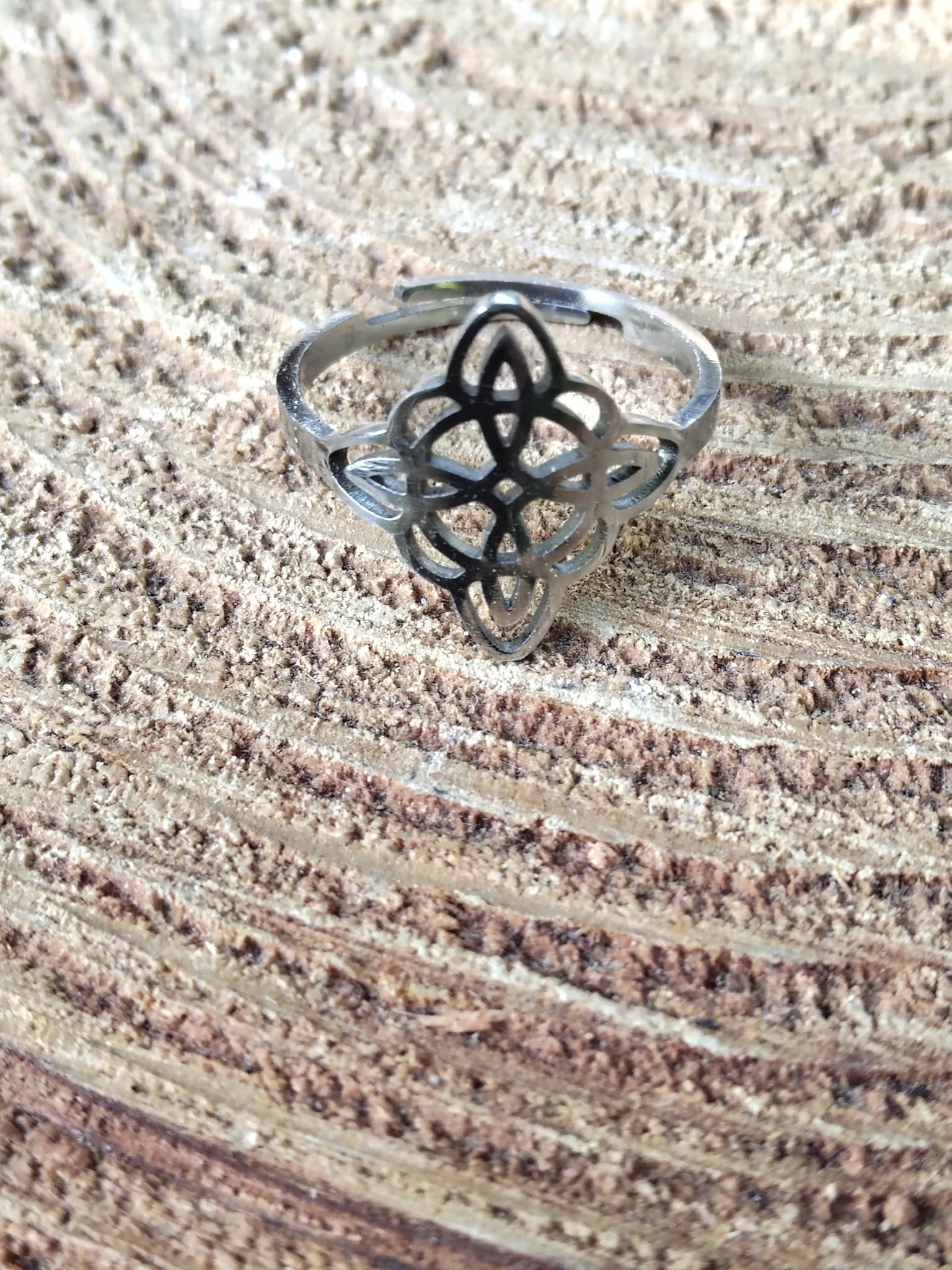 Bague réglable acier inoxydable argenté, nœud de sorcière ajouré, posée sur rondin de bois, ambiance naturelle; symbole de protection.