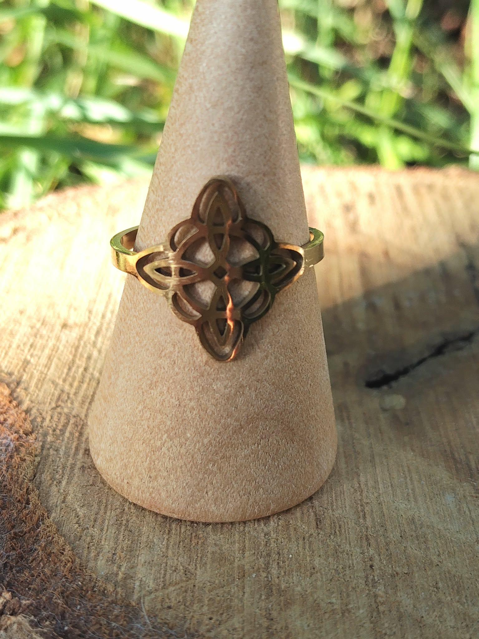 Bague réglable acier inoxydable doré, nœud de sorcière ajouré, sur cône beige, fond végétal flouté; symbole de protection.