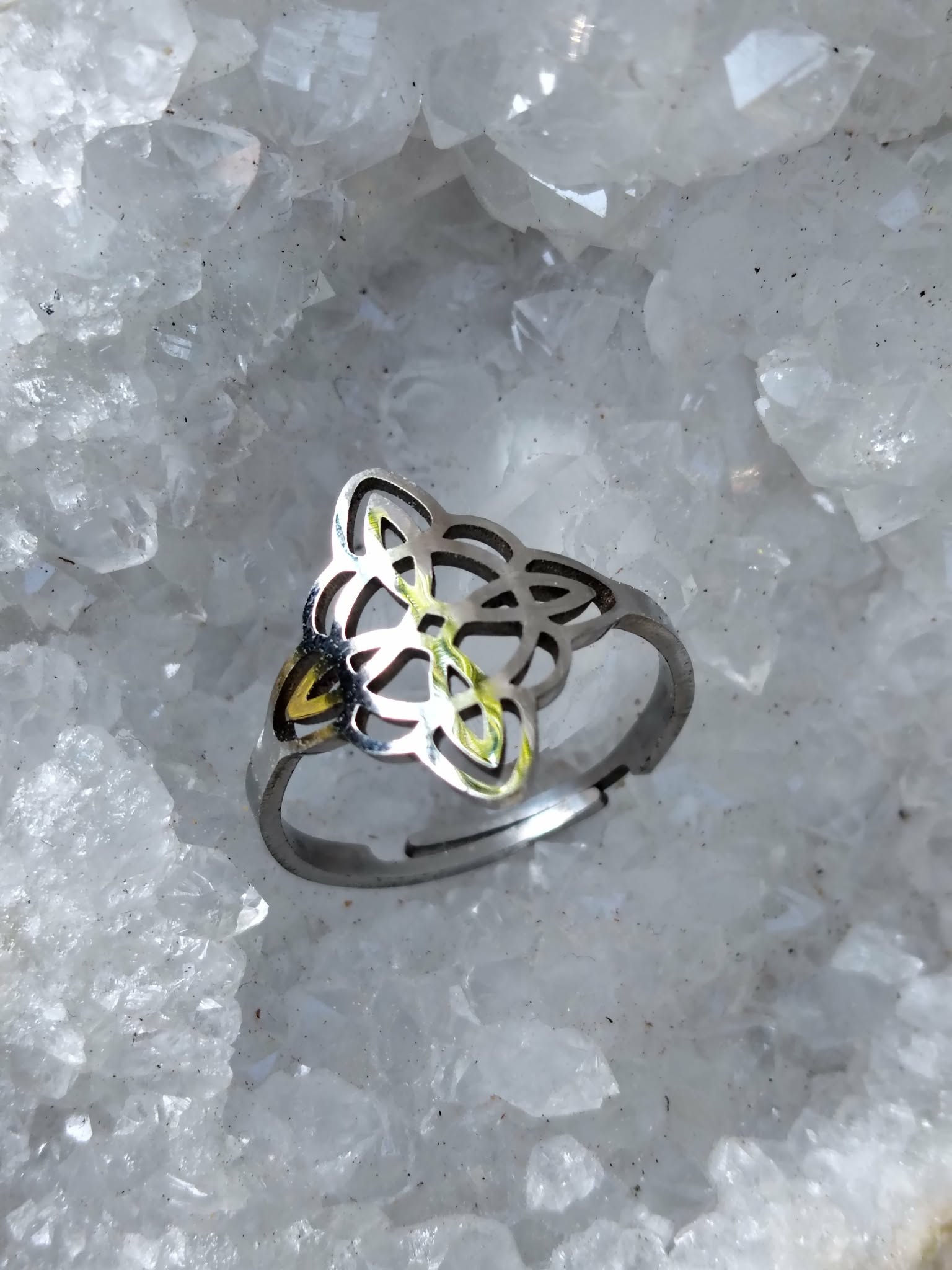 Bague réglable acier inoxydable argenté, nœud de sorcière ajouré, posée sur cristaux blancs (quartz), ambiance minérale; symbole de protection.