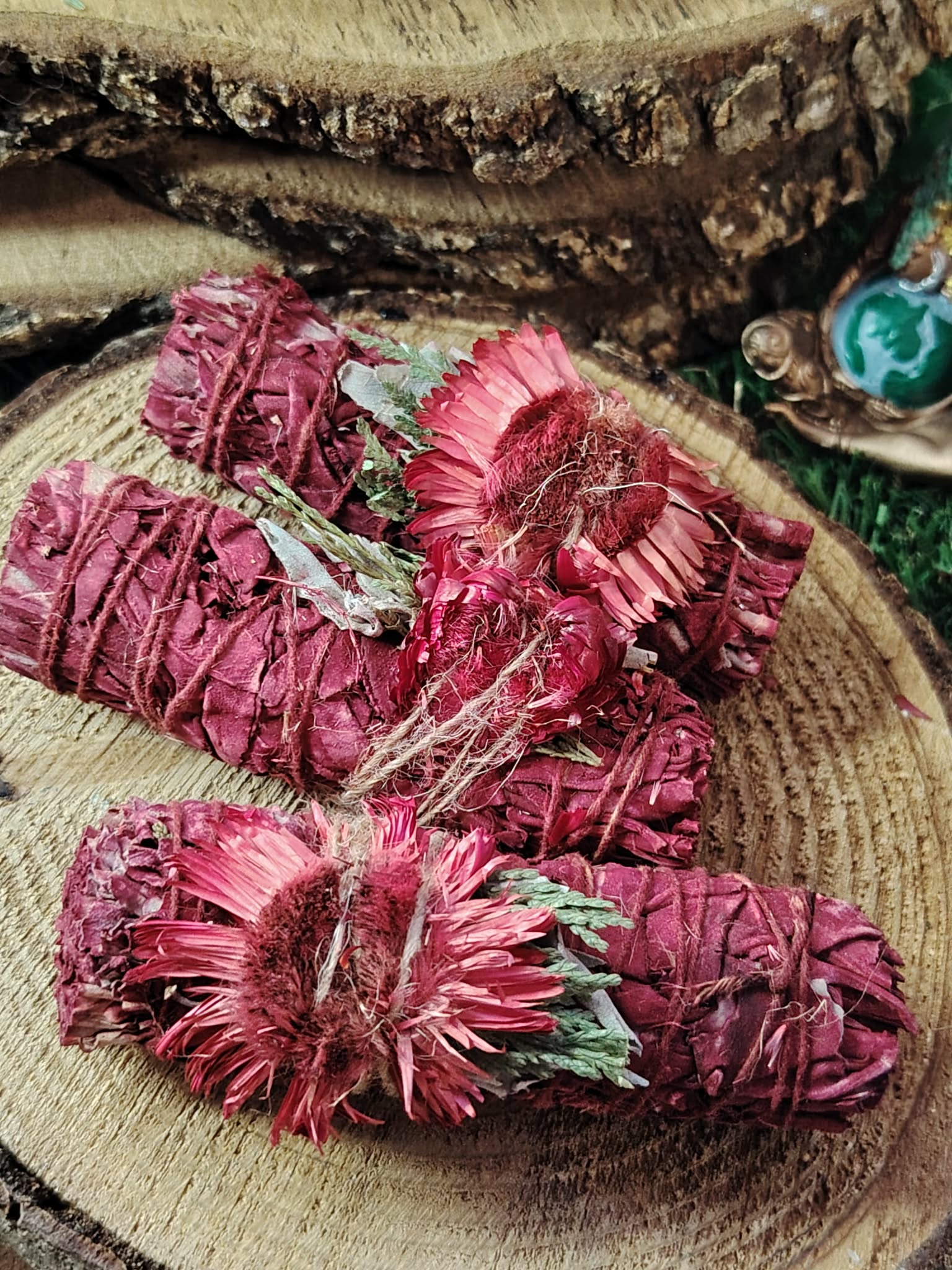 Bâton de fumigation sauge blanche enrobée “sang de dragon”, fleurs séchées roses, ficelle naturelle, posé sur rondin de bois; utilisé pour purifier l’ambiance, poser une intention de protection et de renouveau.