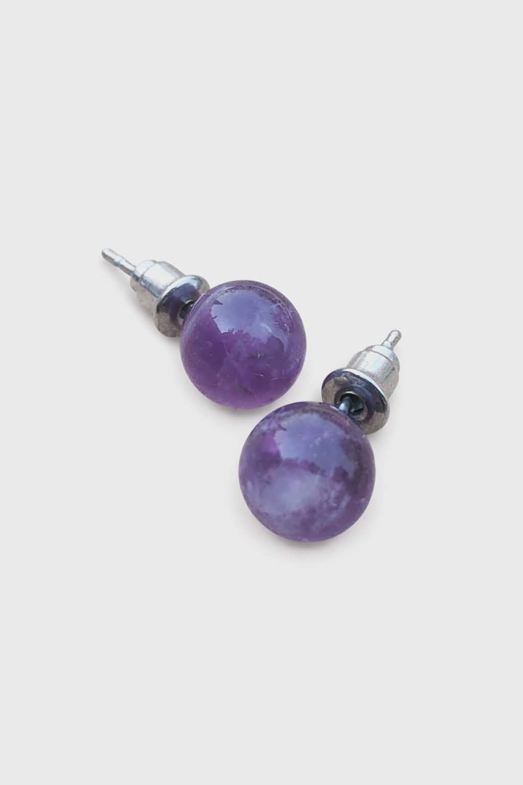 Boucles d’oreilles puces améthyste en gros plan sur fond blanc, perles violettes brillantes, tiges inox 304; associée à sérénité, apaisement, intuition.