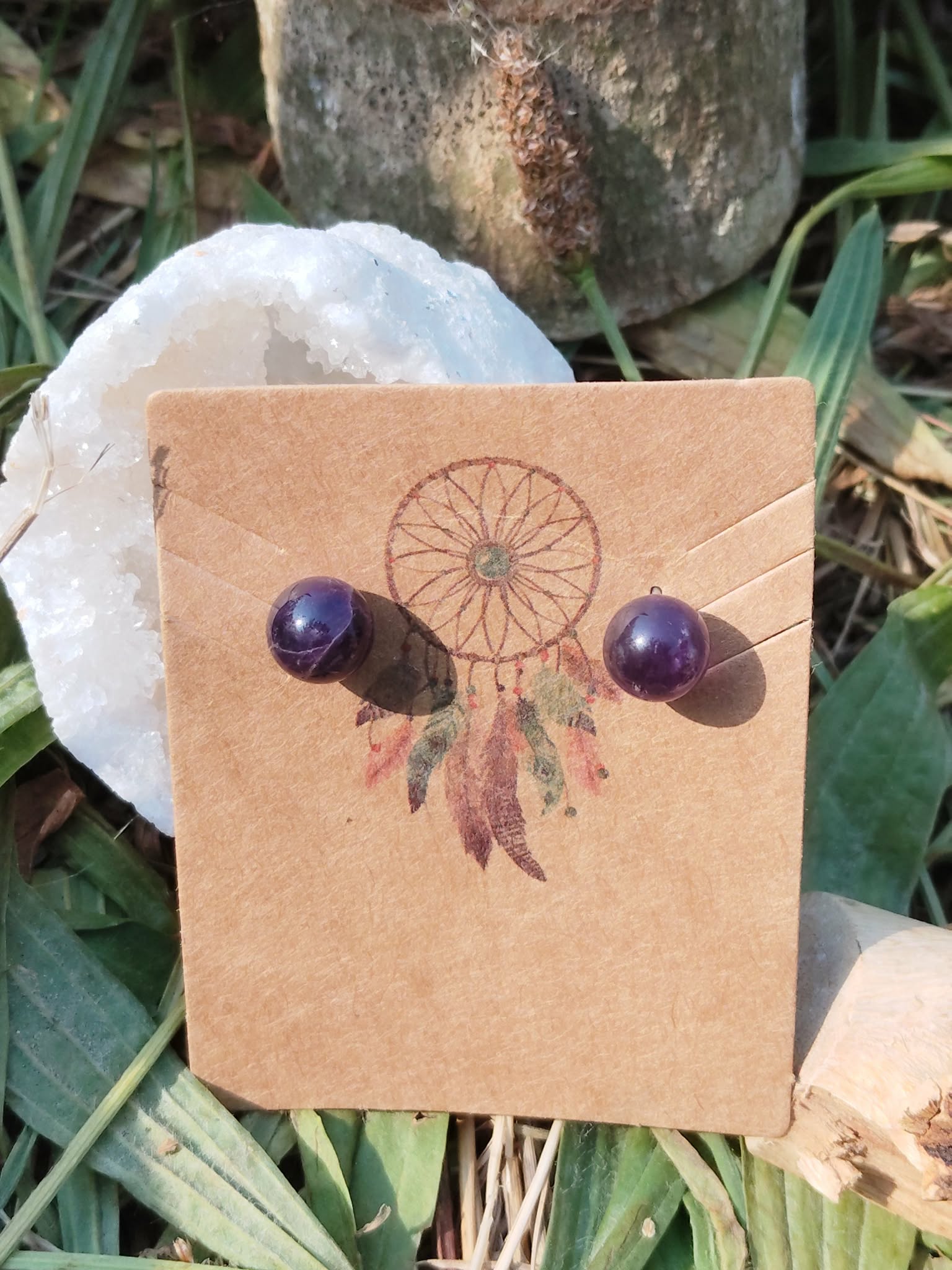 Boucles d’oreilles puces améthyste fixées sur une carte kraft avec motif attrape-rêves, posée devant une coquille blanche, décor d’herbes vertes en arrière-plan; évoque calme, douceur, protection (tradition).