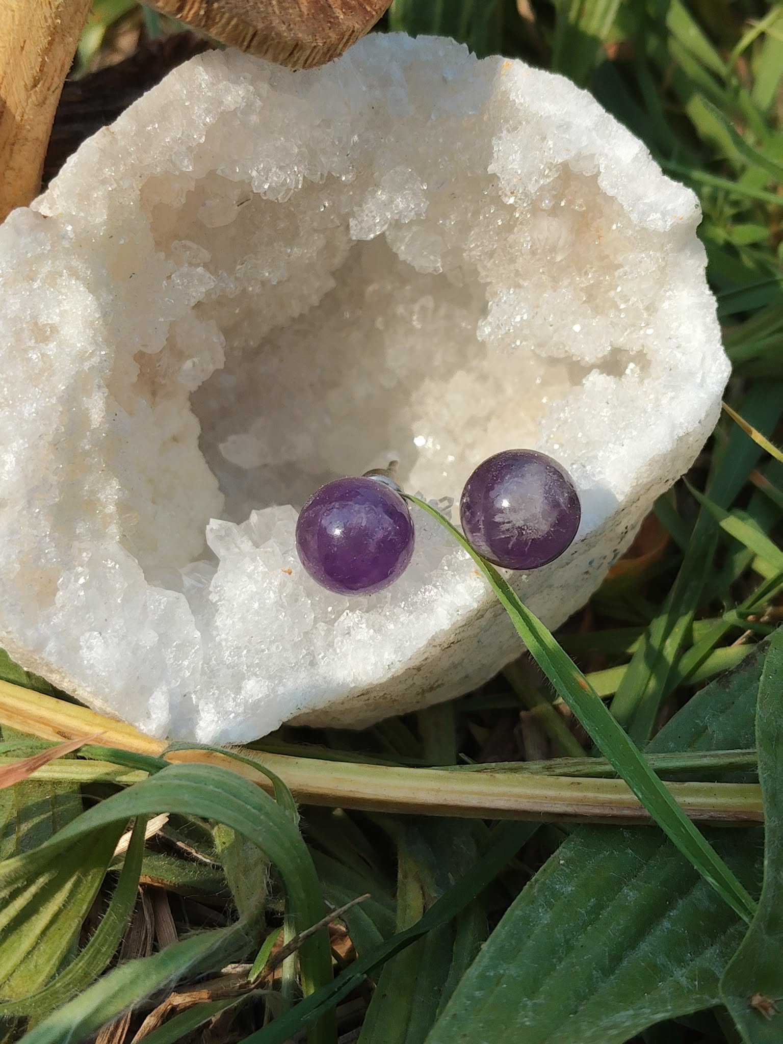 Boucles d’oreilles puces améthyste en duo sur pierre blanche irrégulière, brins verts qui traversent le cadre, photo en extérieur; évoque apaisement, harmonie, douceur.