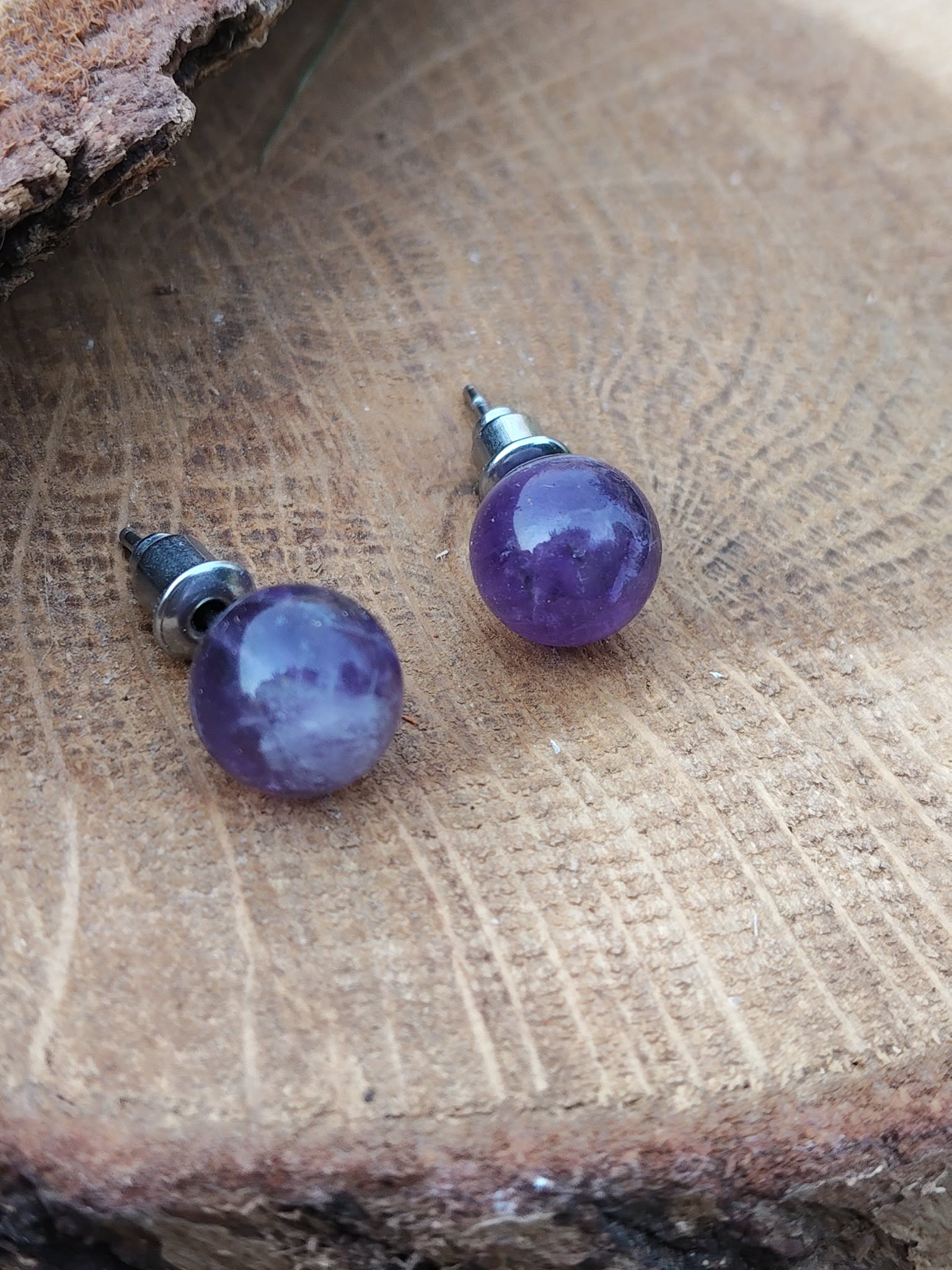 Boucles d’oreilles puces améthyste en duo sur bois brut, reflets sur les perles, arrière-plan neutre; évoque apaisement, harmonie, lâcher-prise.