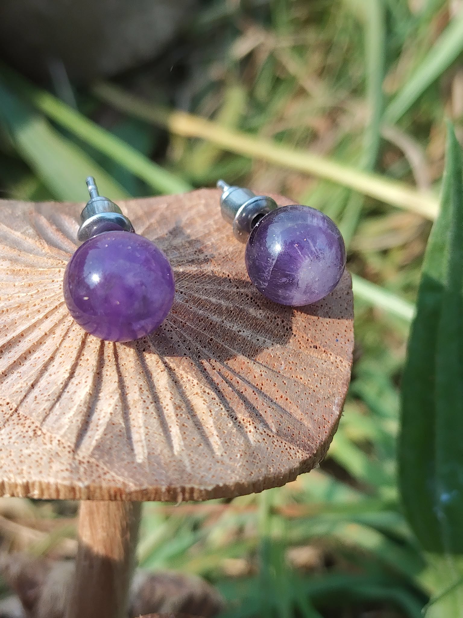 Boucles d’oreilles puces améthyste violette sur tranche de bois brut, soleil rasant et ombres marquées, arrière-plan végétal; associée à calme intérieur, équilibre émotionnel, intuition.