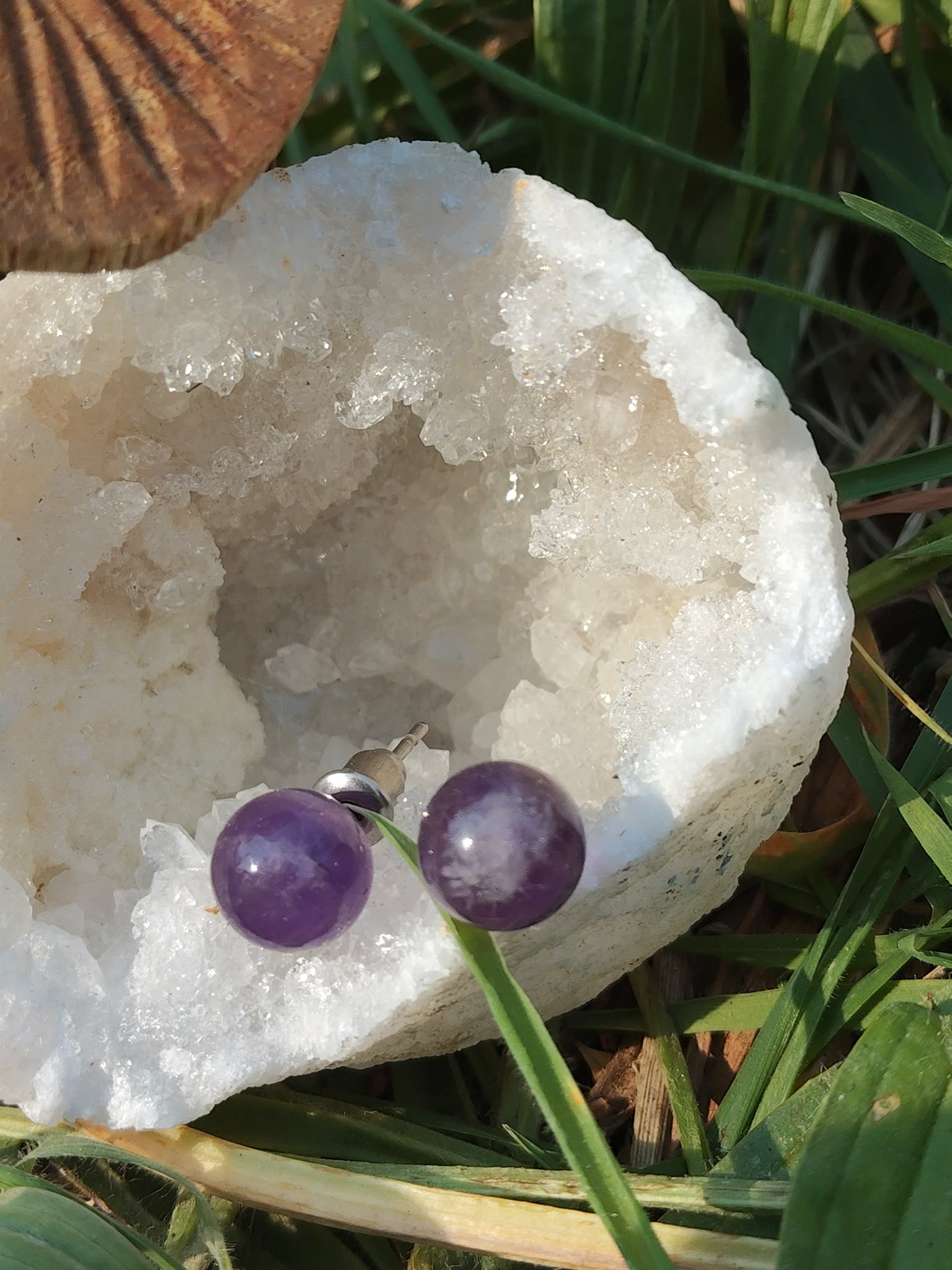 Boucles d’oreilles puces améthyste posées côte à côte sur un amas de quartz, brins d’herbe et teintes naturelles en fond; associée à équilibre émotionnel, paix intérieure, protection (tradition).