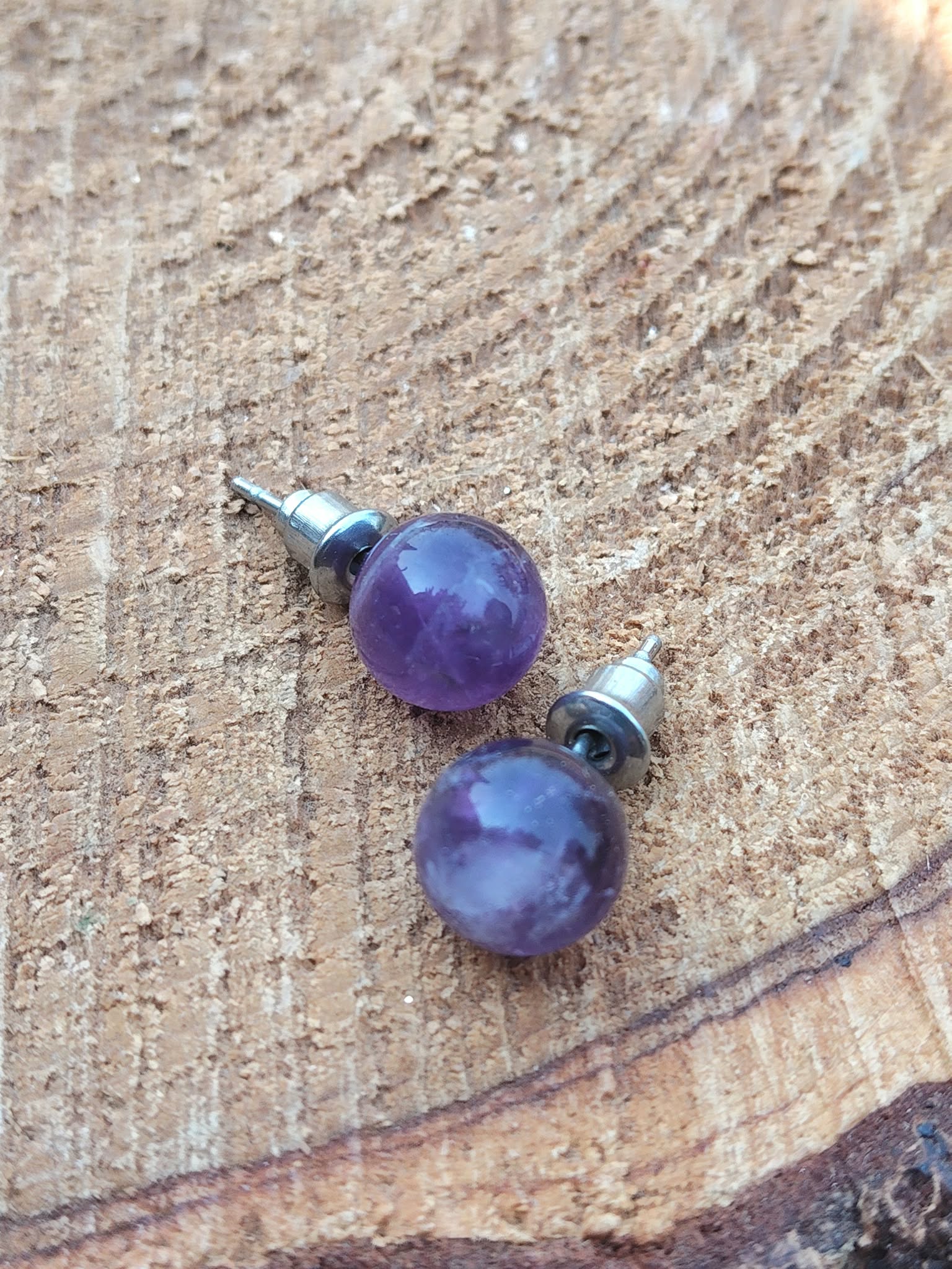 Boucles d’oreilles puces améthyste en inox 304 posées sur bois brut, deux perles violettes avec reflets, lumière naturelle; évoque calme, recentrage, clarté.