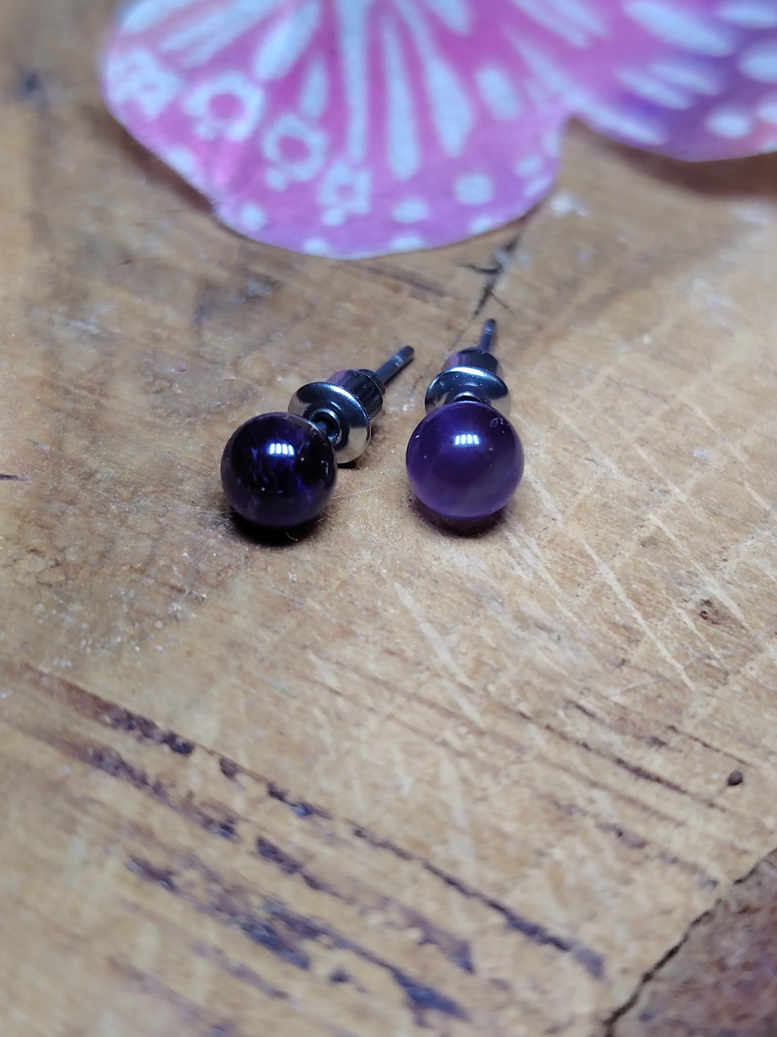 Boucles d’oreilles puces améthyste sur planche en bois, duo de perles violettes en premier plan, papillon rose en arrière-plan légèrement flouté; associée à sérénité, douceur, intuition.