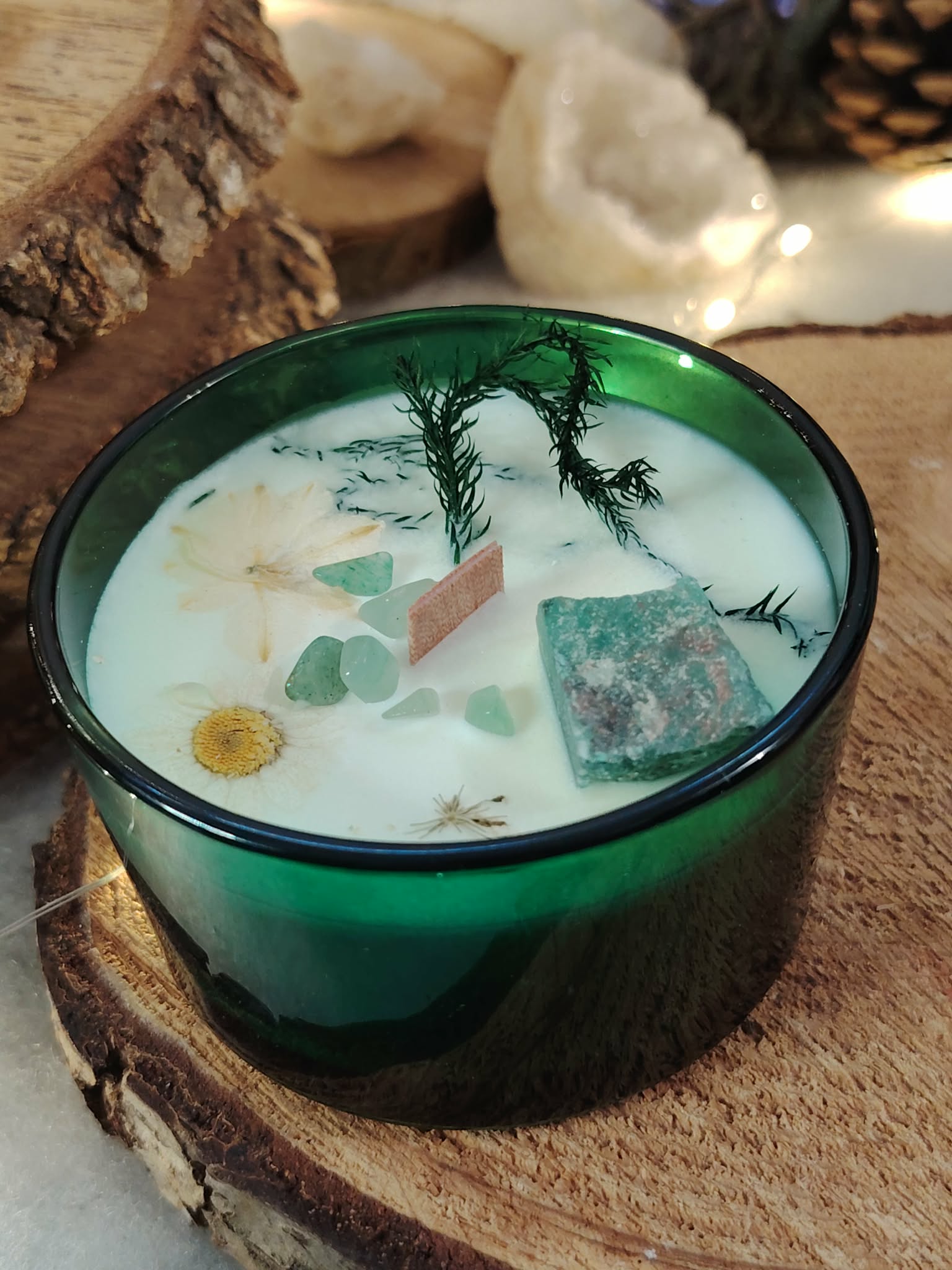 Bougie jade menthe-citron en cire de soja, pot en verre vert; cire blanche avec éclats de jade et fleurs séchées, posée sur rondin de bois, fond clair.