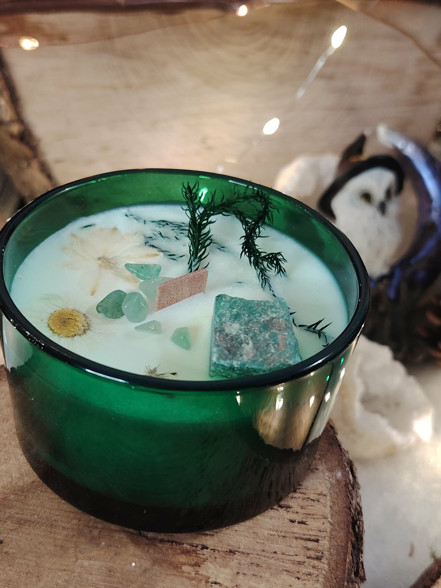 Gros plan bougie “jade” menthe-citron en cire de soja; verre vert, cire blanche ornée de pierres vertes, décor cocooning avec guirlande et pierre claire à côté.