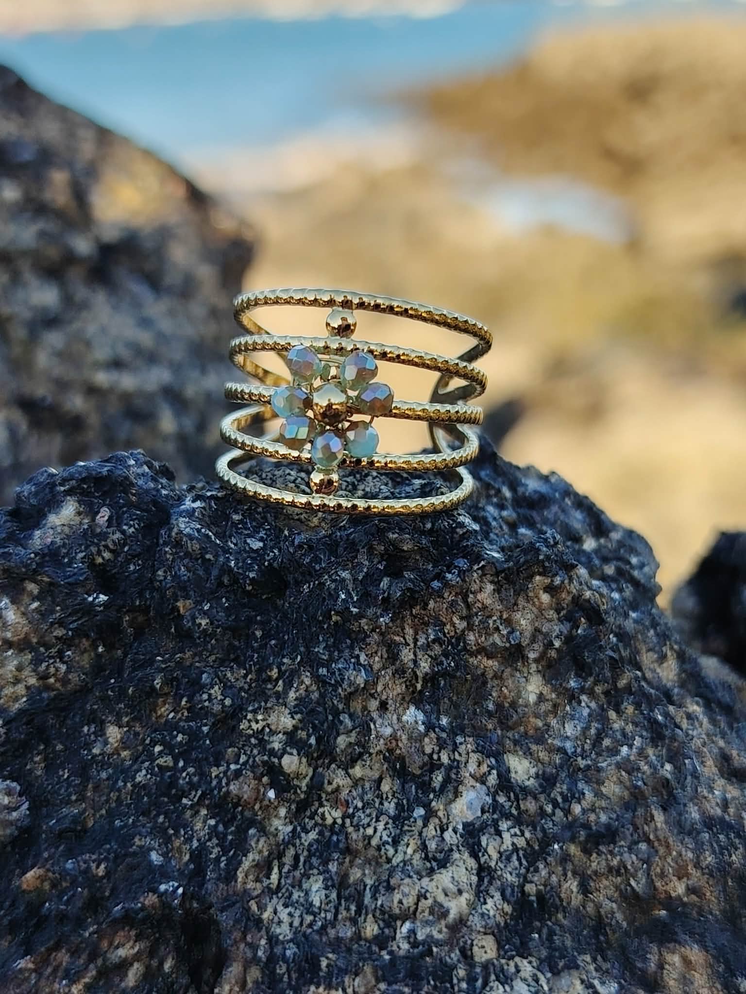 Bague dorée multi-anneaux torsadés en acier inoxydableavec cristaux bleus irisés, posée sur un rocher en bord de mer, arrière-plan plage flouté, photo prise en Bretagne.