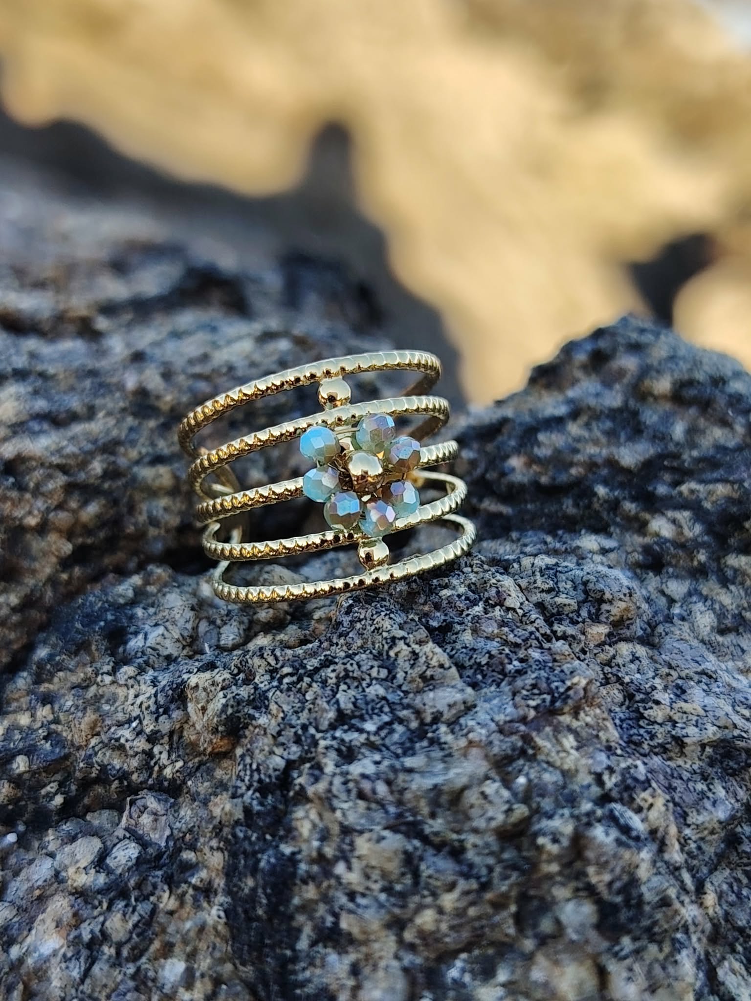 Bague dorée acier inoxydable style spirale multi anneaux, grappe de cristaux bleus au centre, mise en scène sur rocher granitique, fond mer et plage en flou artistique, photo en Bretagne.