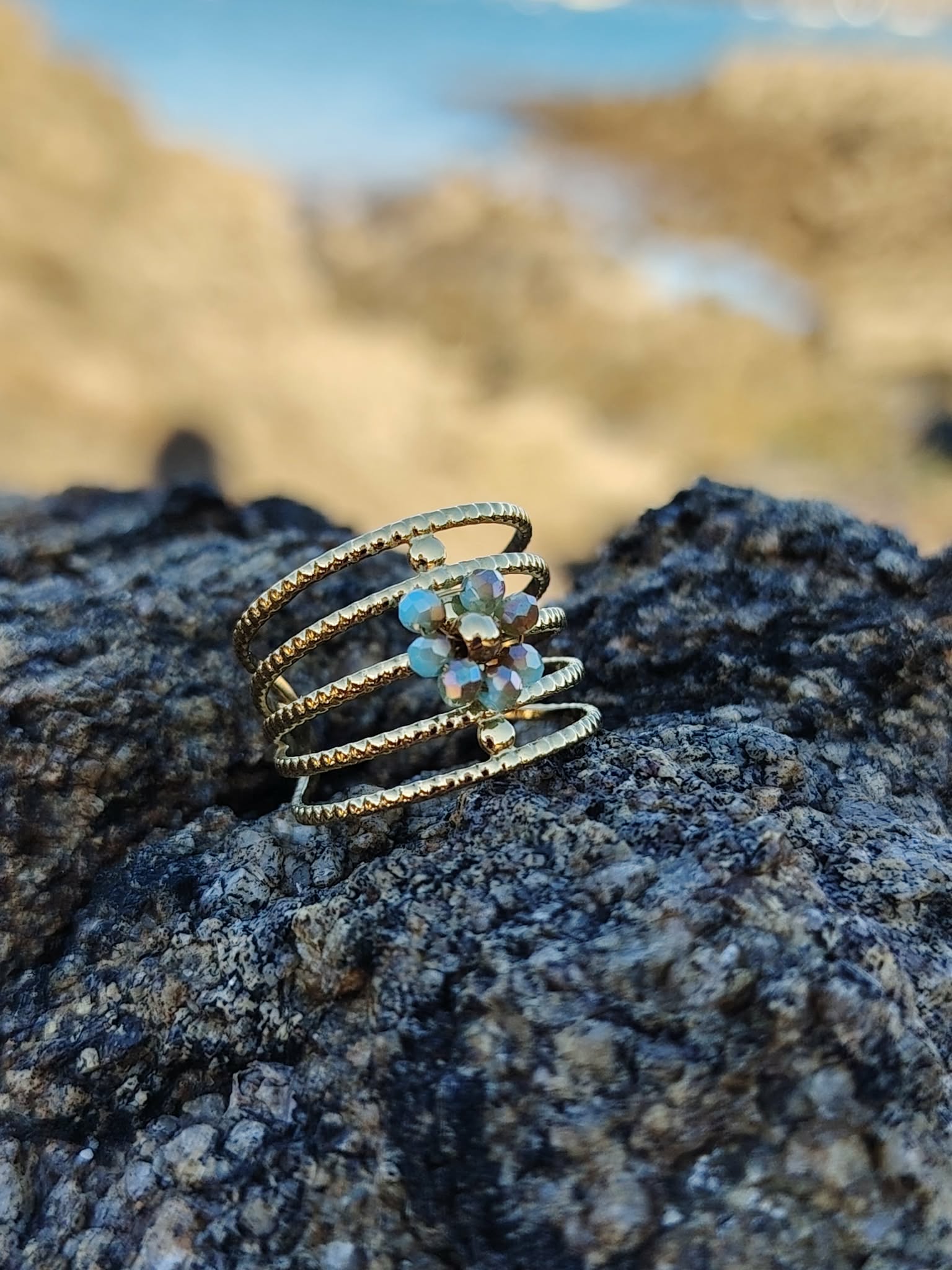 Bague dorée multi-anneaux torsadés acier inoxydable avec cristaux bleus, posée sur rochers sombres en bord de mer, plage et eau floutées en arrière-plan, photo prise en Bretagne.