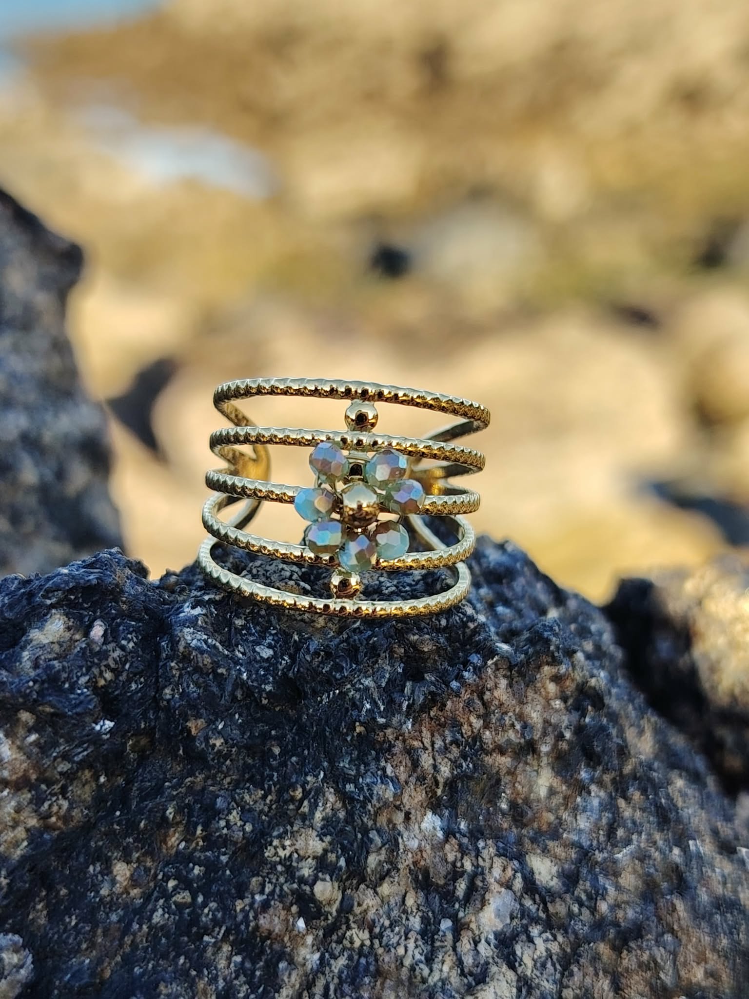 Bague dorée à plusieurs rangs texturés en acier inoxydable, ornée de petits cristaux bleus irisés, mise en scène sur rocher noir en bord de mer, fond sable flou, photo en Bretagne.