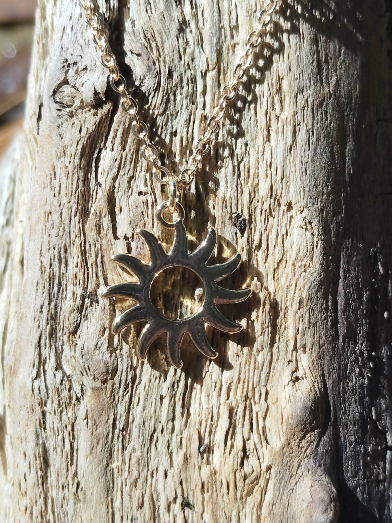Pendentif soleil doré en acier inoxydable sur chaîne fine, photographié sur bois patiné en Bretagne, style lumineux et présence chaleureuse