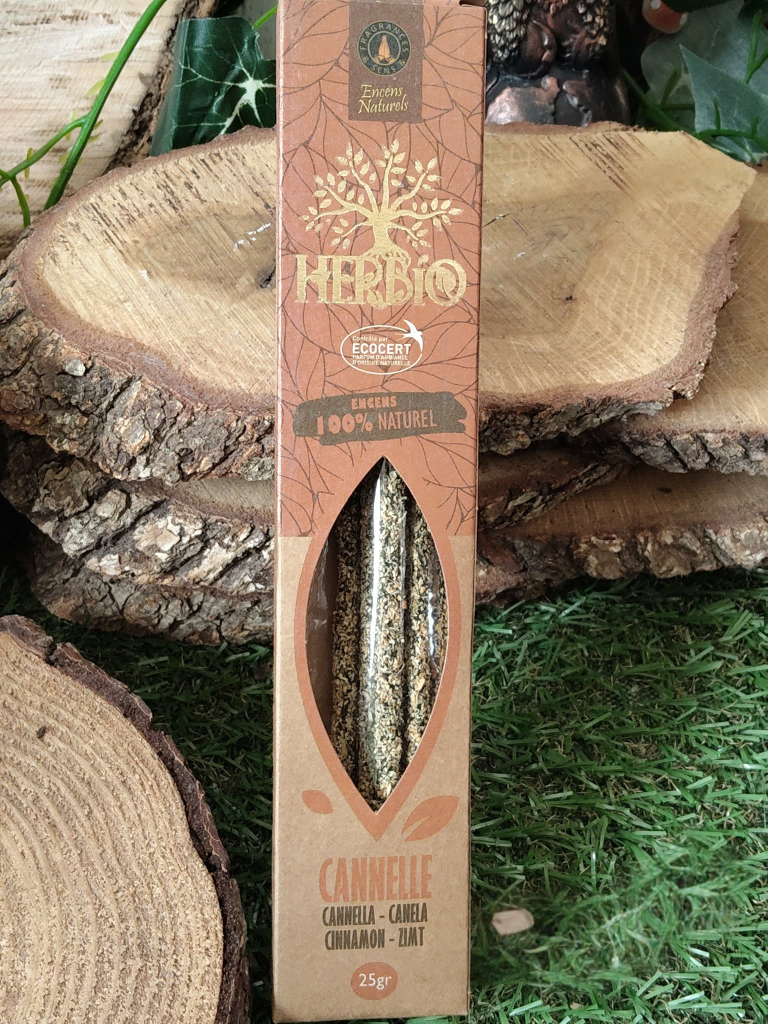 Encens HERBIO Cannelle 25 g en étui kraft avec fenêtre, présenté devant des rondins de bois et verdure; senteur épicée, ambiance chaleureuse et réconfortante.