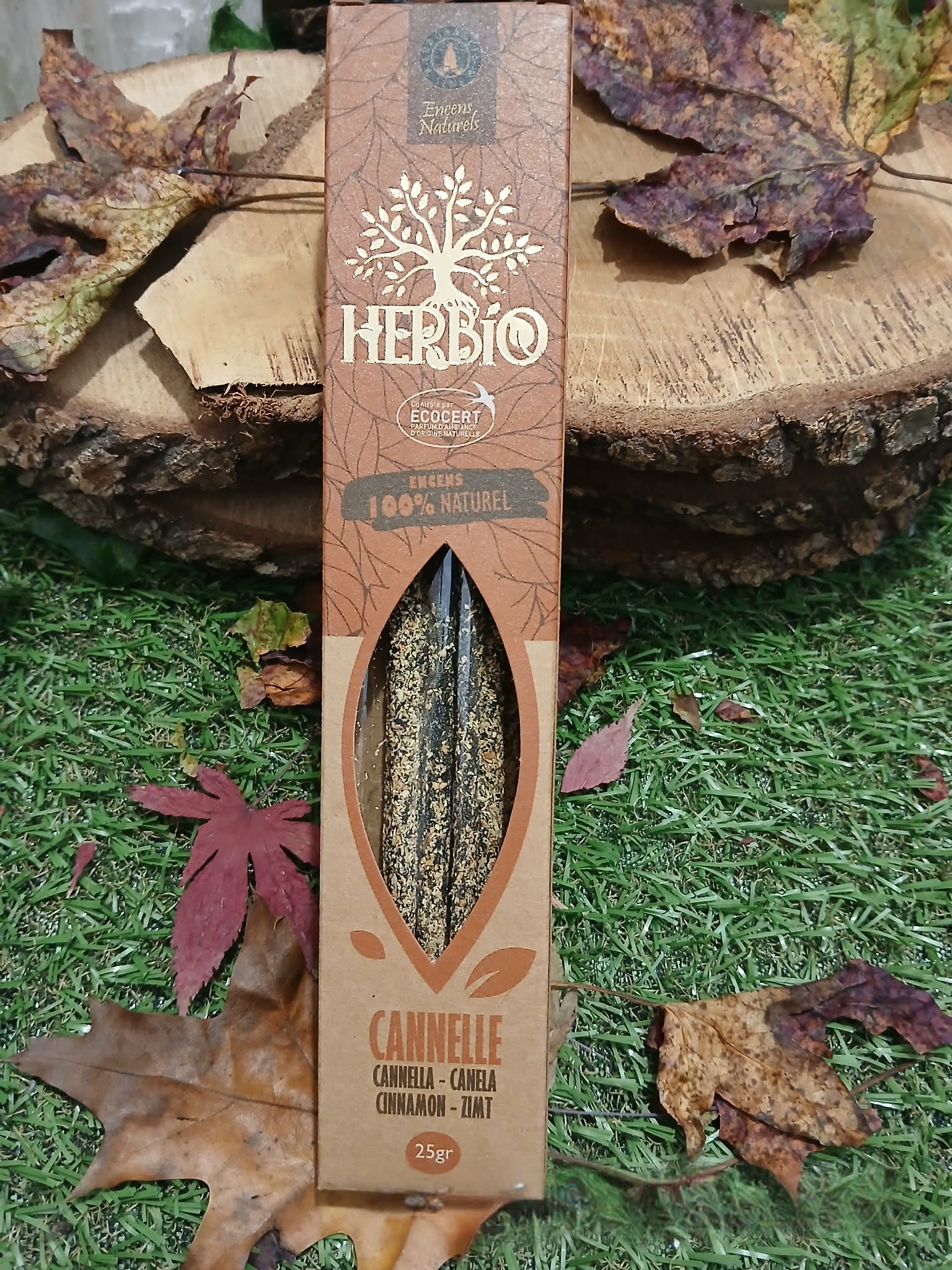 Encens HERBIO Cannelle 25 g en étui kraft avec fenêtre, mis en scène sur herbe et feuilles d’automne avec rondin de bois; note épicée, atmosphère réconfortante et conviviale