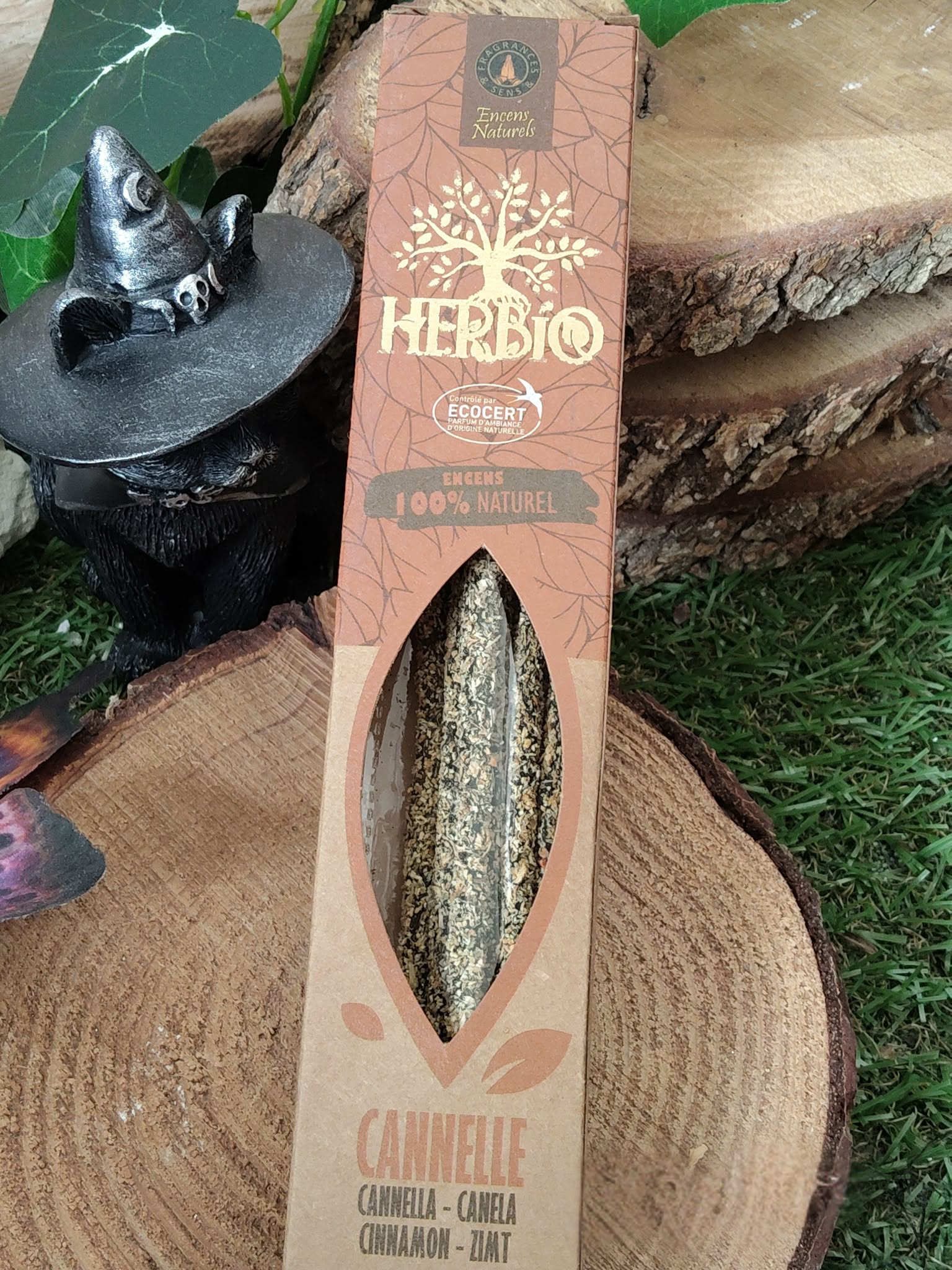 Encens HERBIO Cannelle 25 g, étui kraft avec fenêtre, sur tranche de bois en décor automnal (feuilles, herbe) avec petite figurine argentée; ambiance chaleureuse et réconfortante.