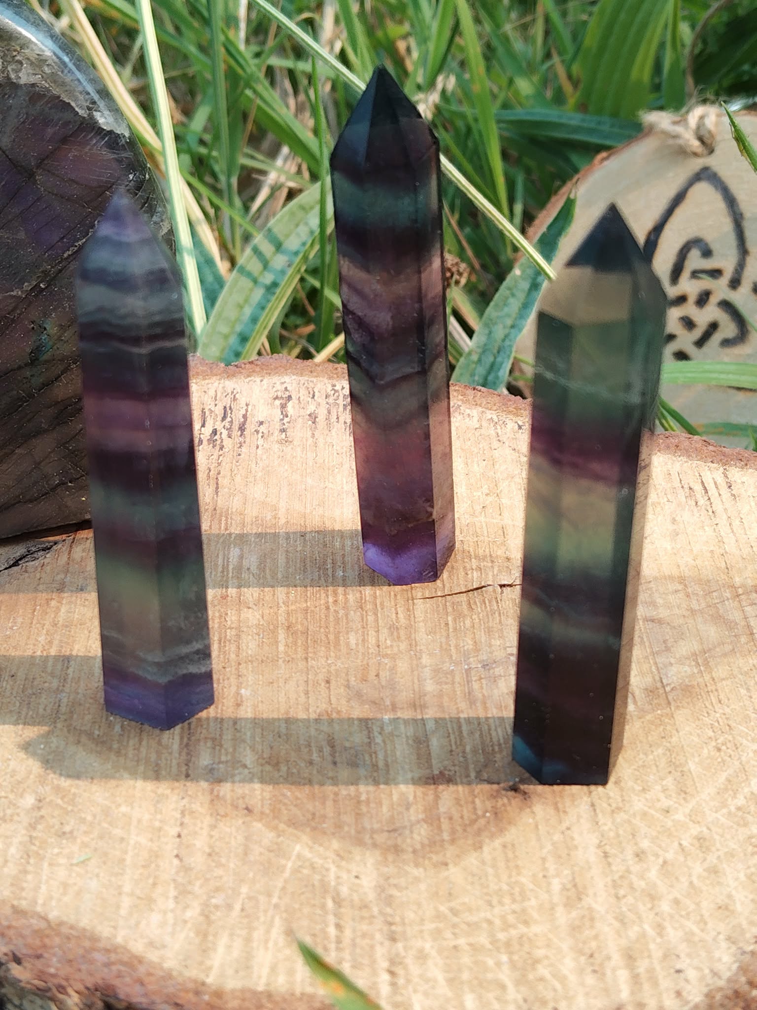 Pointe de fluorite multicolore aux nuances vertes et violettes, posée sur support en bois avec herbes en arrière-plan, pierre naturelle associée à la lucidité, au recul et à l’harmonie intérieure.