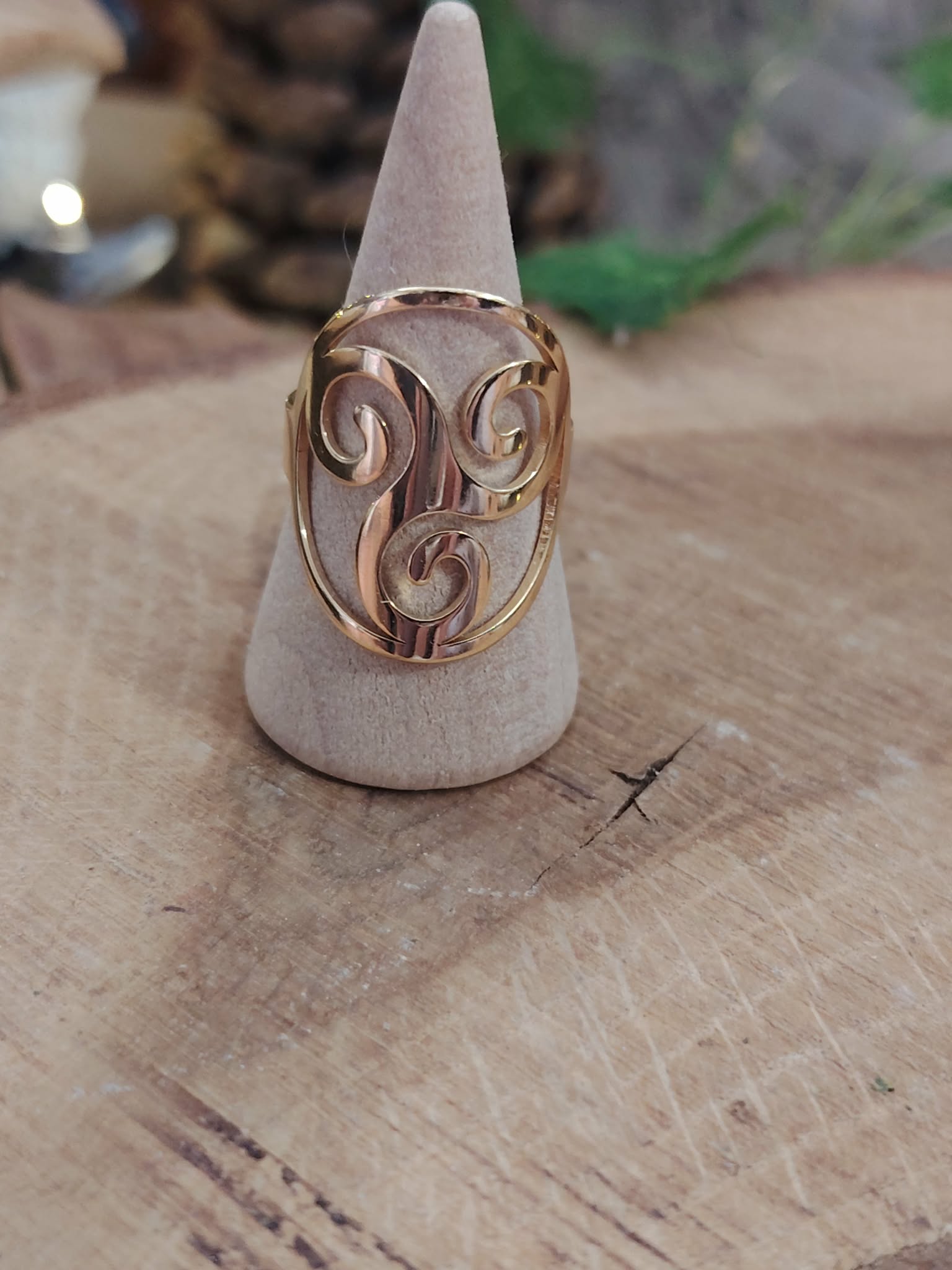 Bague Triskell dorée réglable en acier inoxydable sur porte-bague conique blanc, posée sur planche en bois; symbole cycles, équilibre, protection.