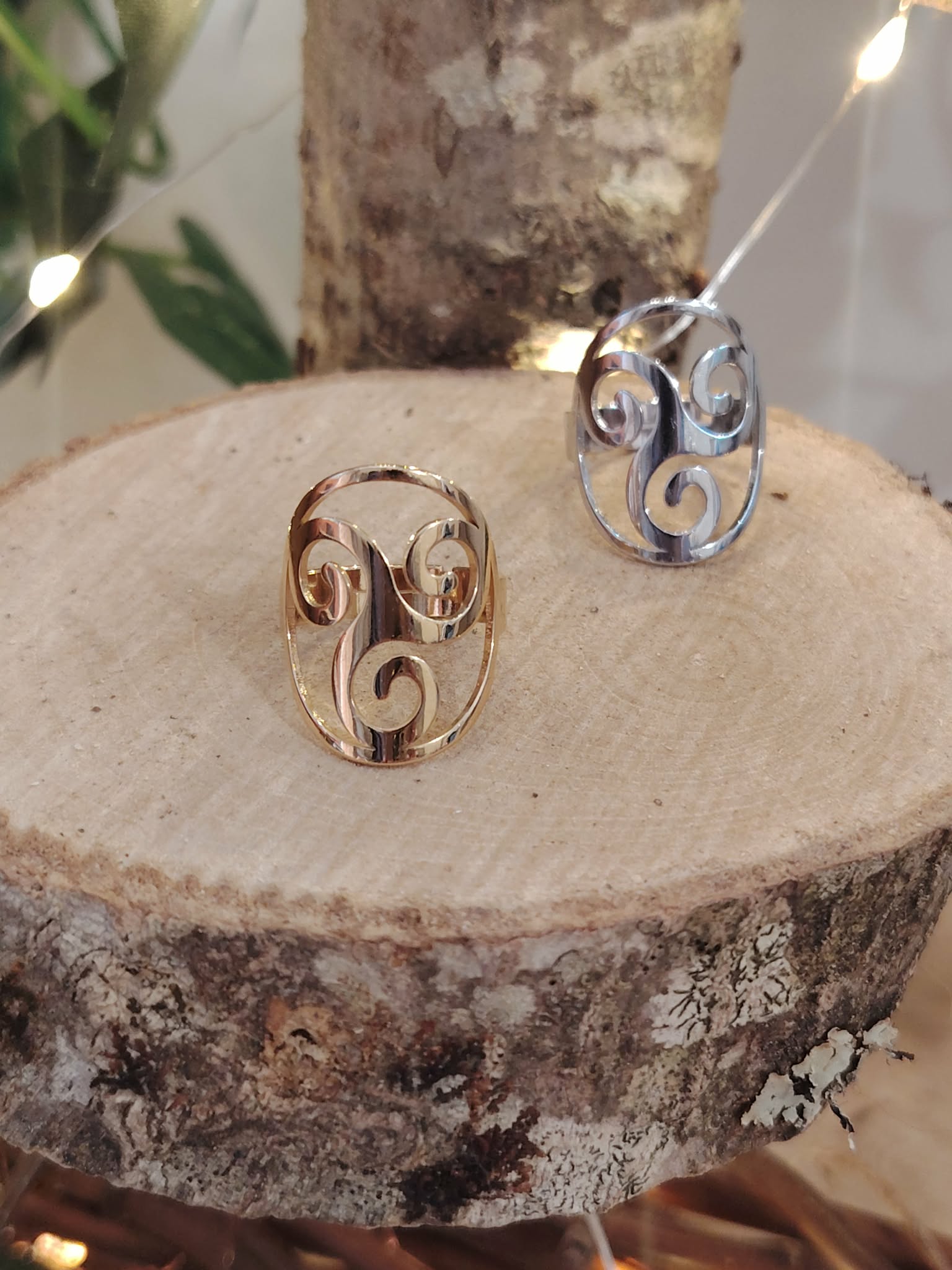 Bague Triskell dorée et bague Triskell argentée posées sur rondin de bois, feuillage en fond; symbole cycles et équilibre.
