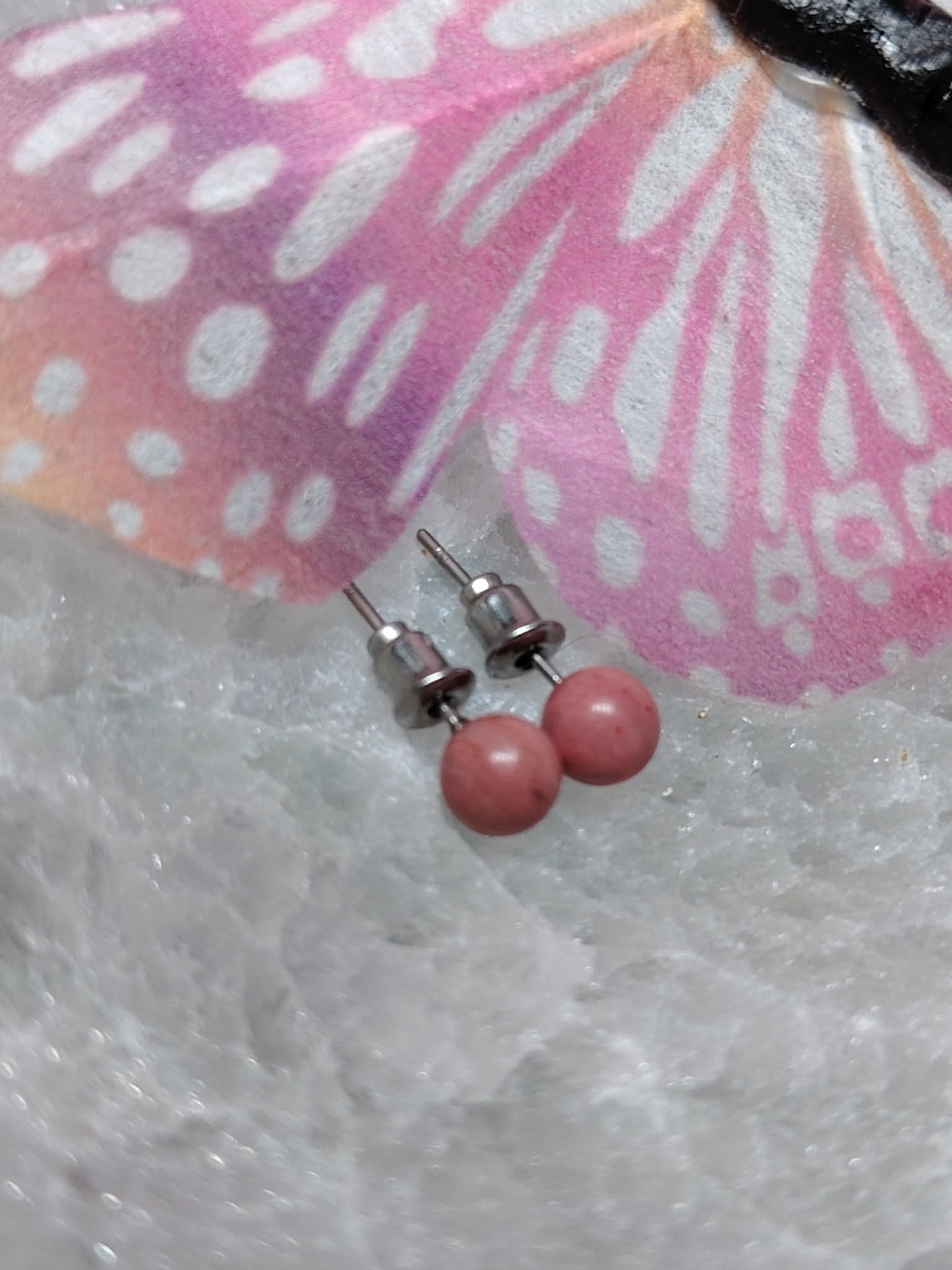 Boucles d’oreilles puces cornaline orange, tiges acier inoxydable, posées sur géode claire avec papillon rose en décor; énergie, vitalité et confiance.